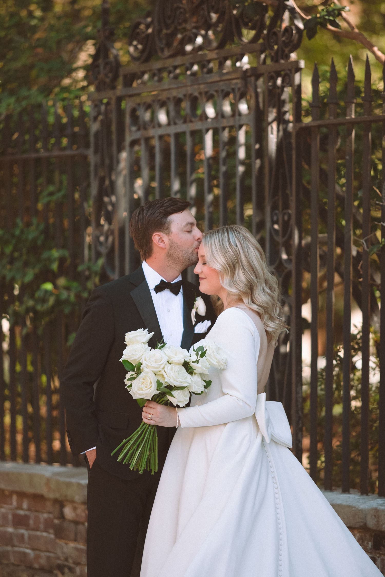 Groom in black tuxedo kissing bride in white gown holding white rose bouquet by ornate iron gate.