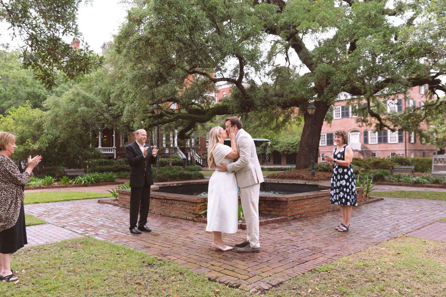 Couple shares first kiss at outdoor wedding ceremony in a brick courtyard with large oak trees and small fountain.