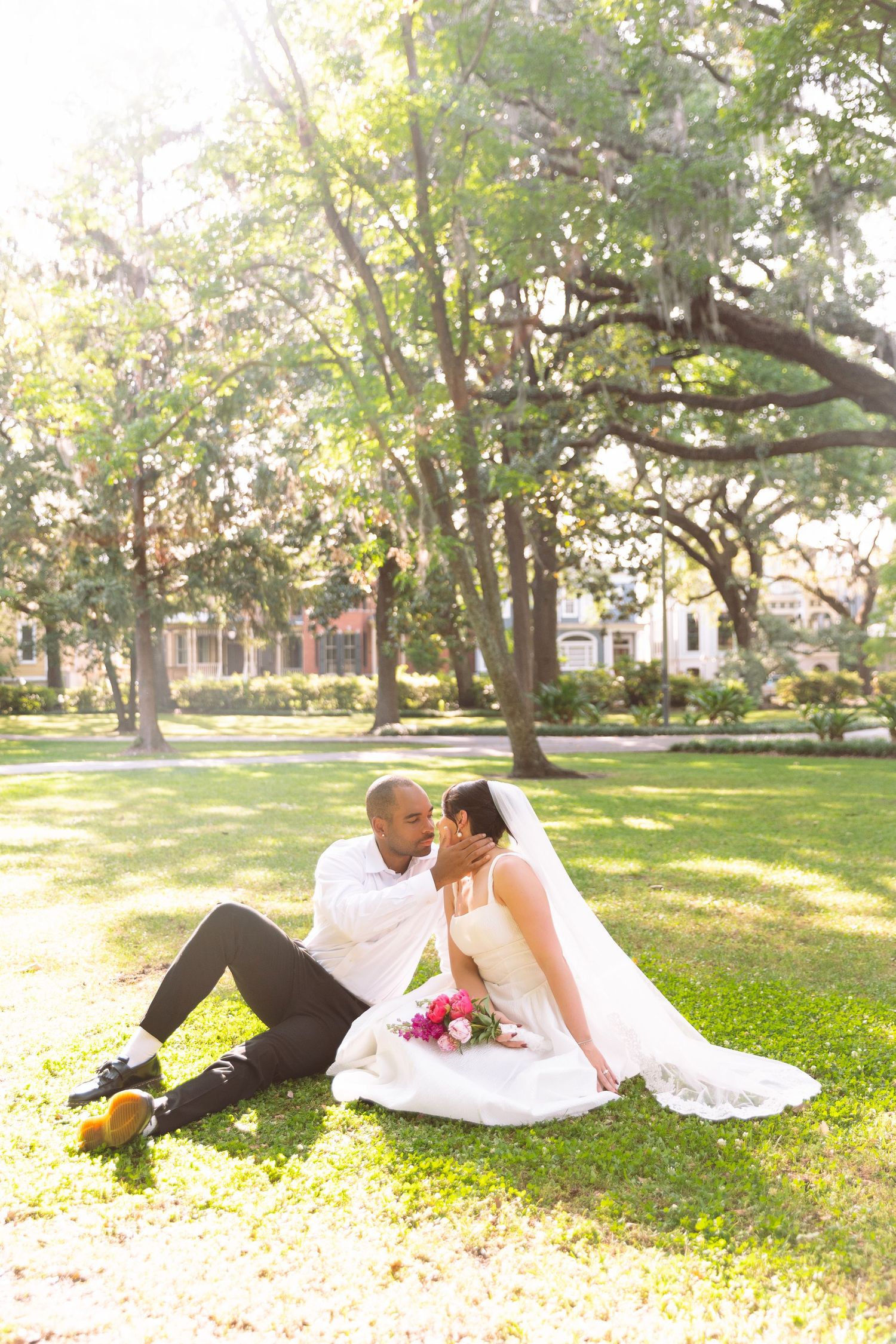 Newlywed couple sitting on green grass in a sunlit park, sharing a tender kiss on their wedding day.