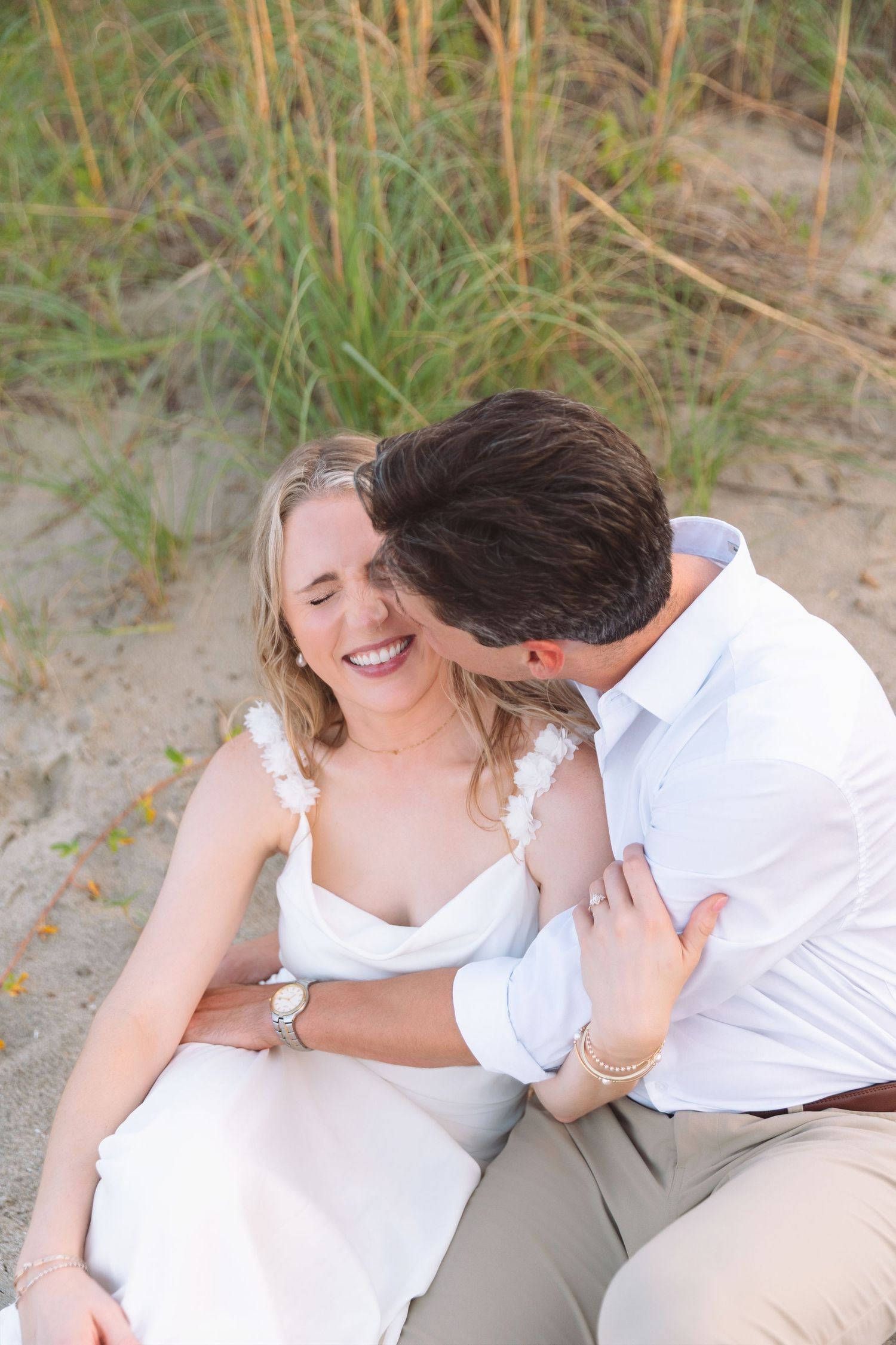 Couple sitting on sand dunes, man kissing smiling woman near tall beach grass.