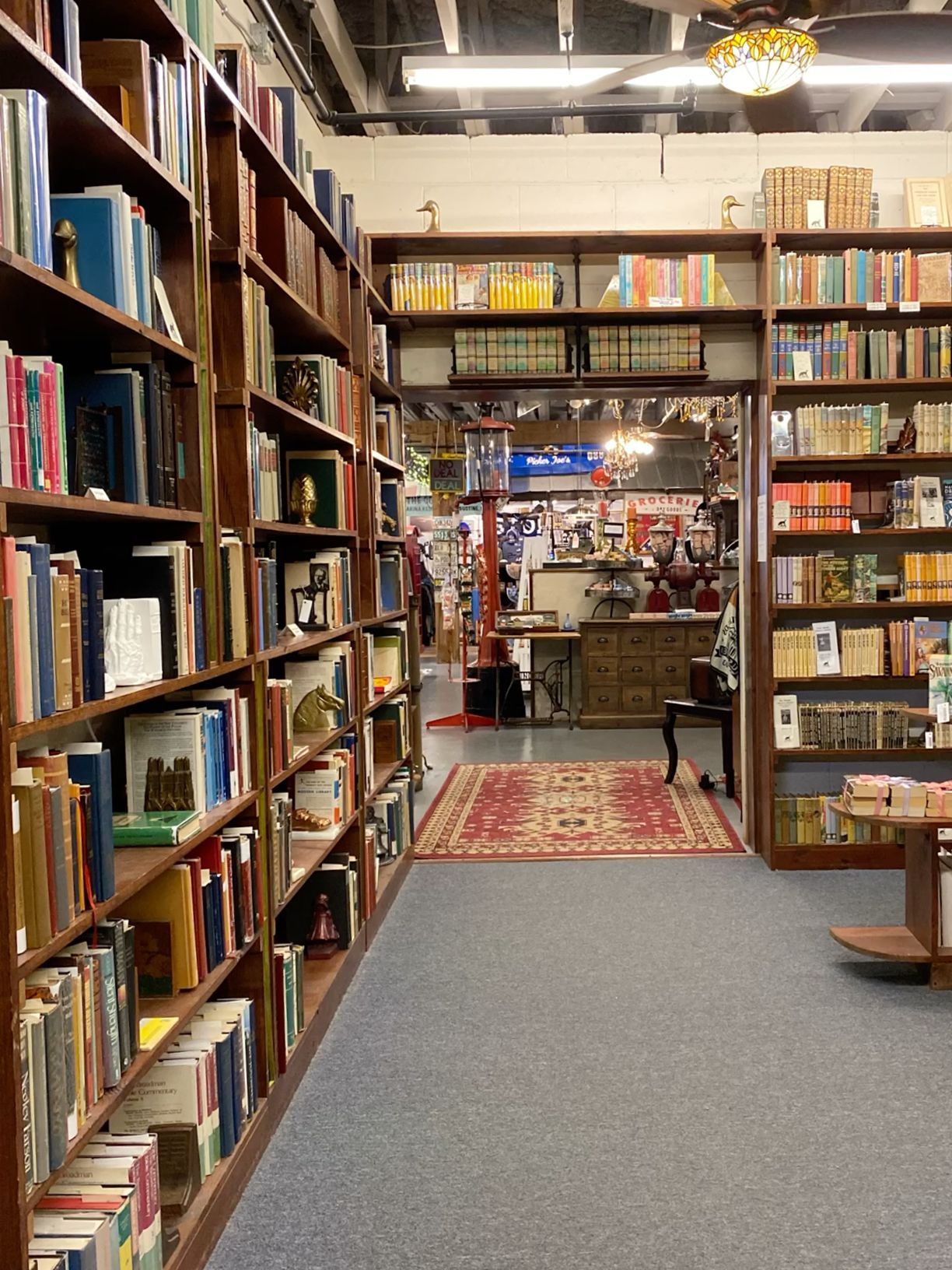Cozy used bookstore aisle with tall wooden shelves lined with books and a red patterned rug on the floor.