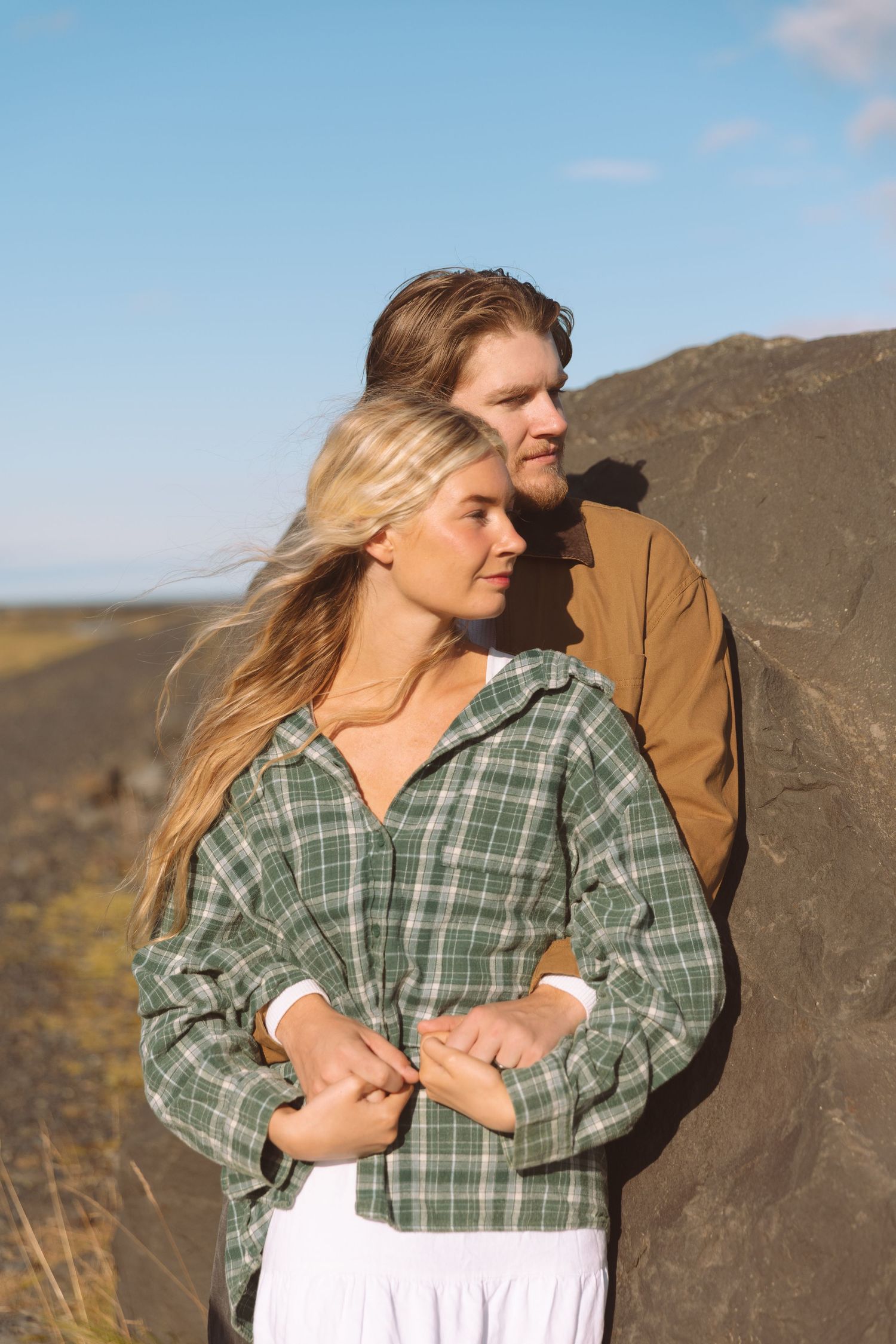 Man embraces woman from behind as they lean against a large rock in open landscape.