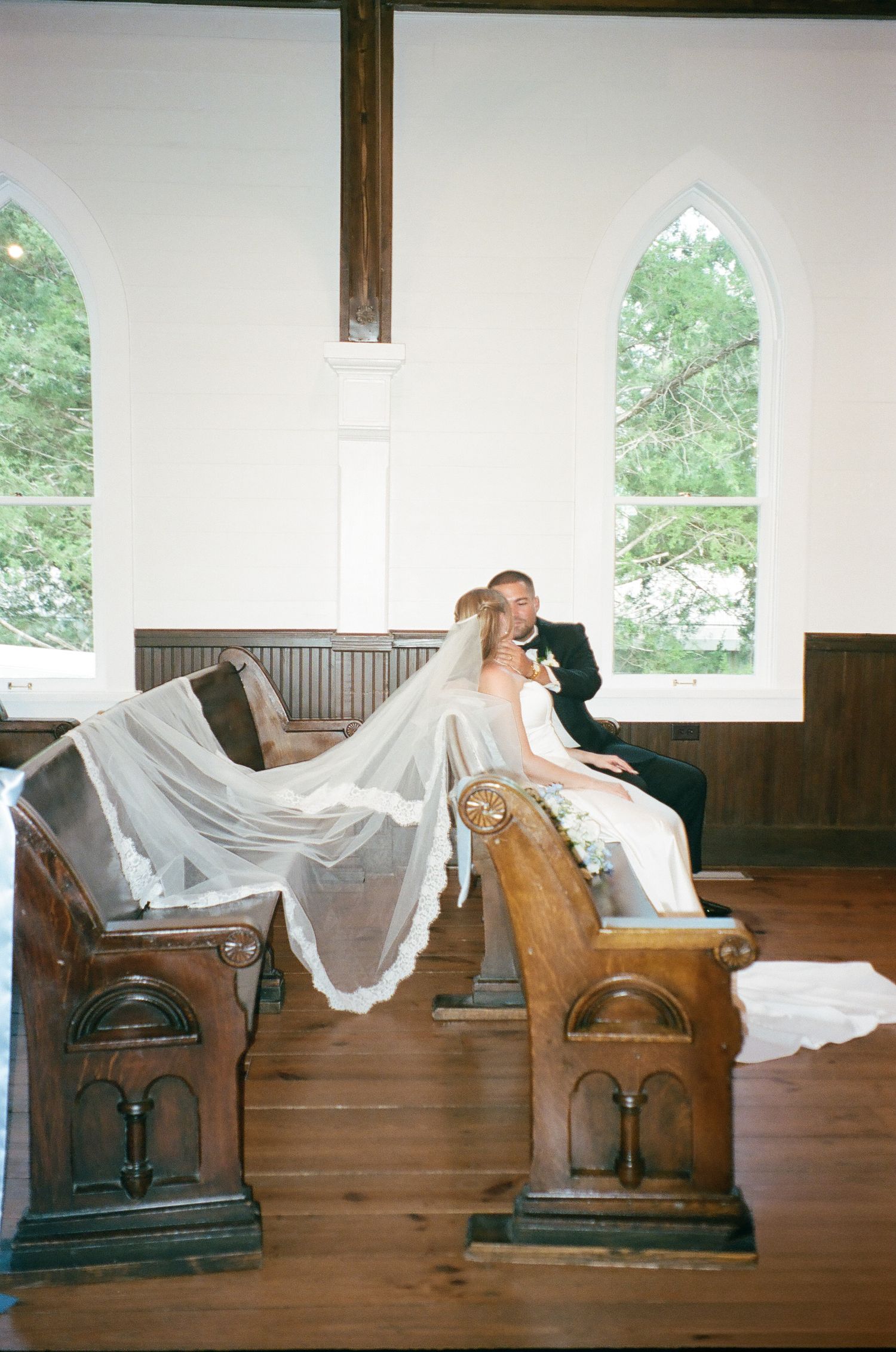 Bride and groom share a romantic moment on wooden church pews, with a flowing veil draped over the bench.