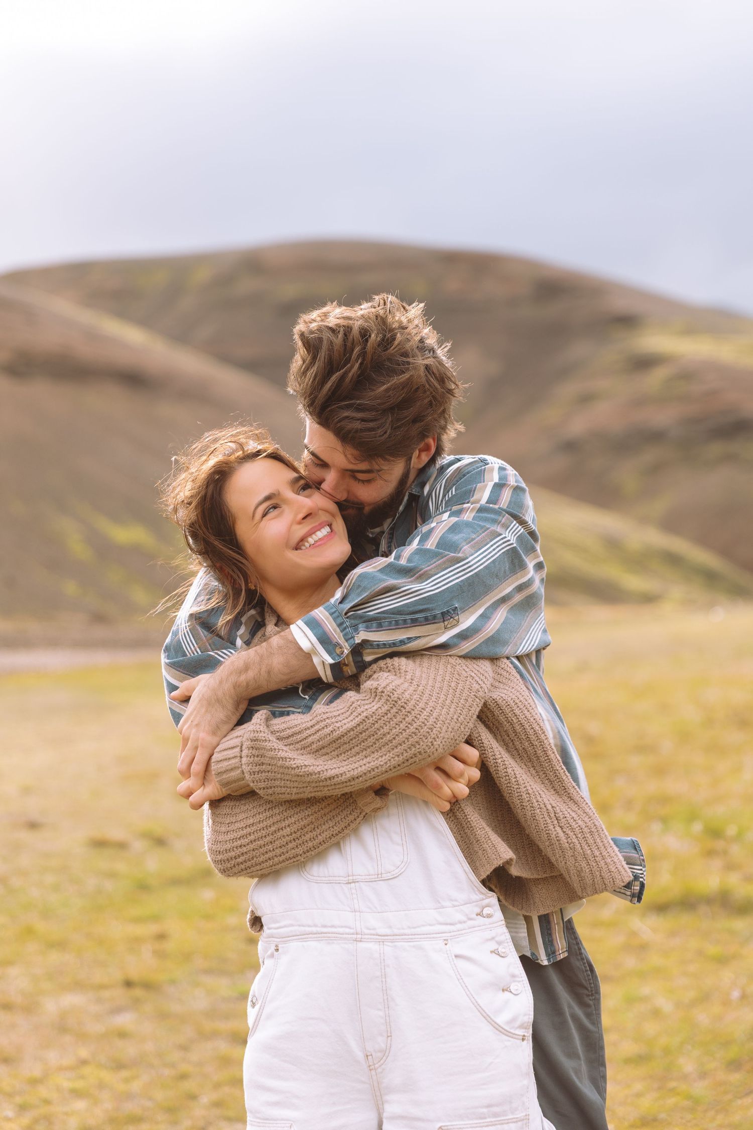 Couple embracing and kissing in a green mountain landscape, woman smiling in white overalls and beige sweater.