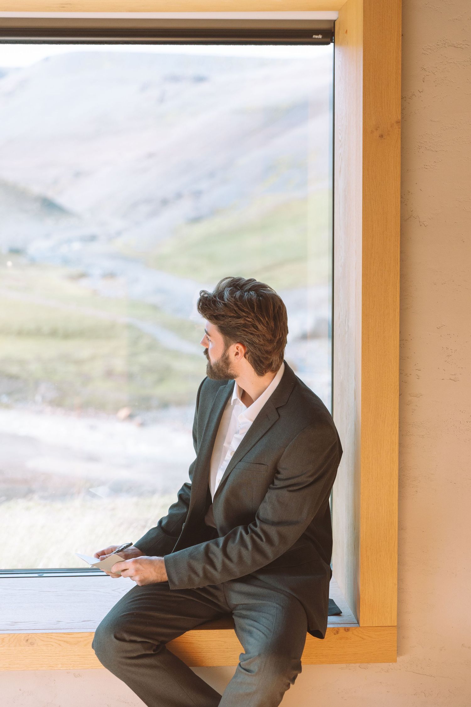 A bearded groom in a dark suit sits by a large window holding paper, gazing at mountain scenery.