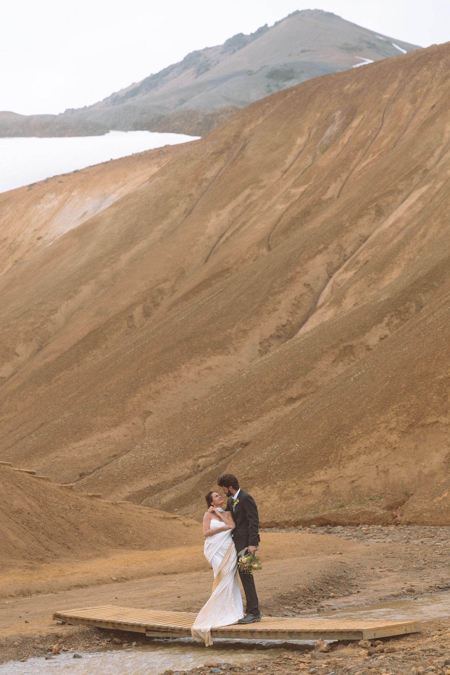 Bride and groom share a romantic embrace on a wooden platform amid Iceland's majestic volcanic mountains.