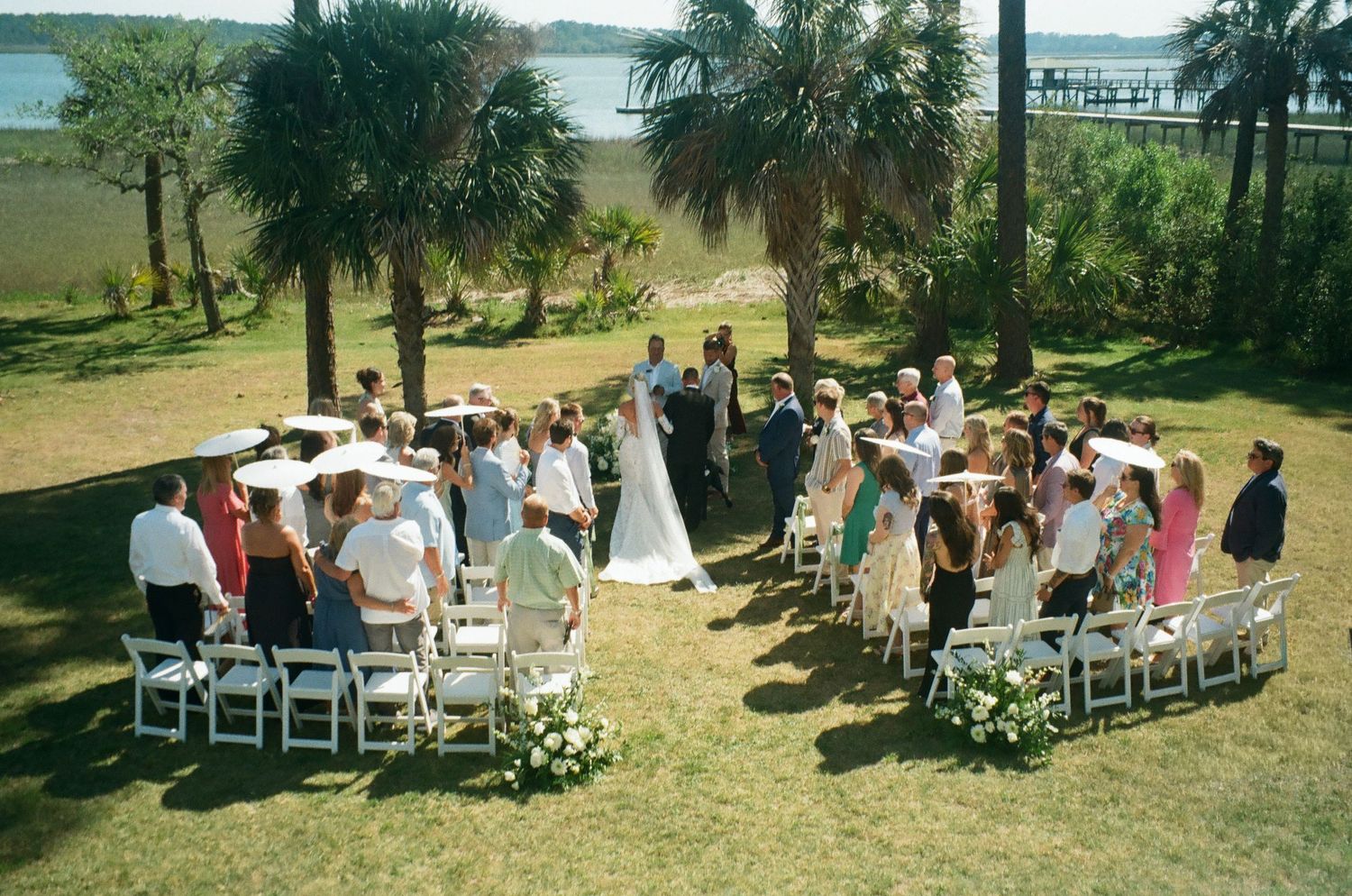 Outdoor wedding ceremony on green lawn with white chairs, palm trees, and waterfront backdrop on a sunny day.
