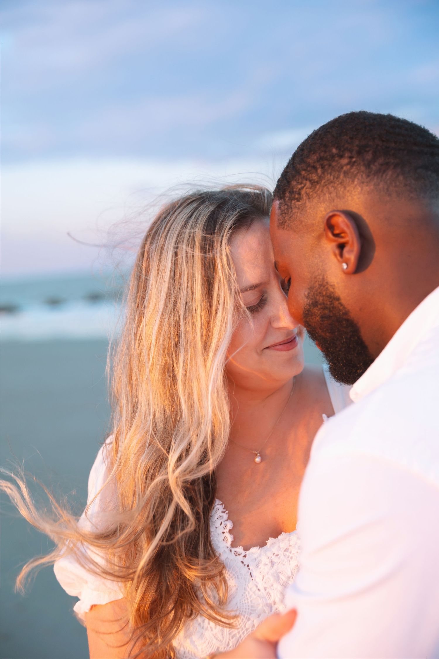 Romantic interracial couple embracing on beach at sunset, woman in white lace dress with eyes closed.