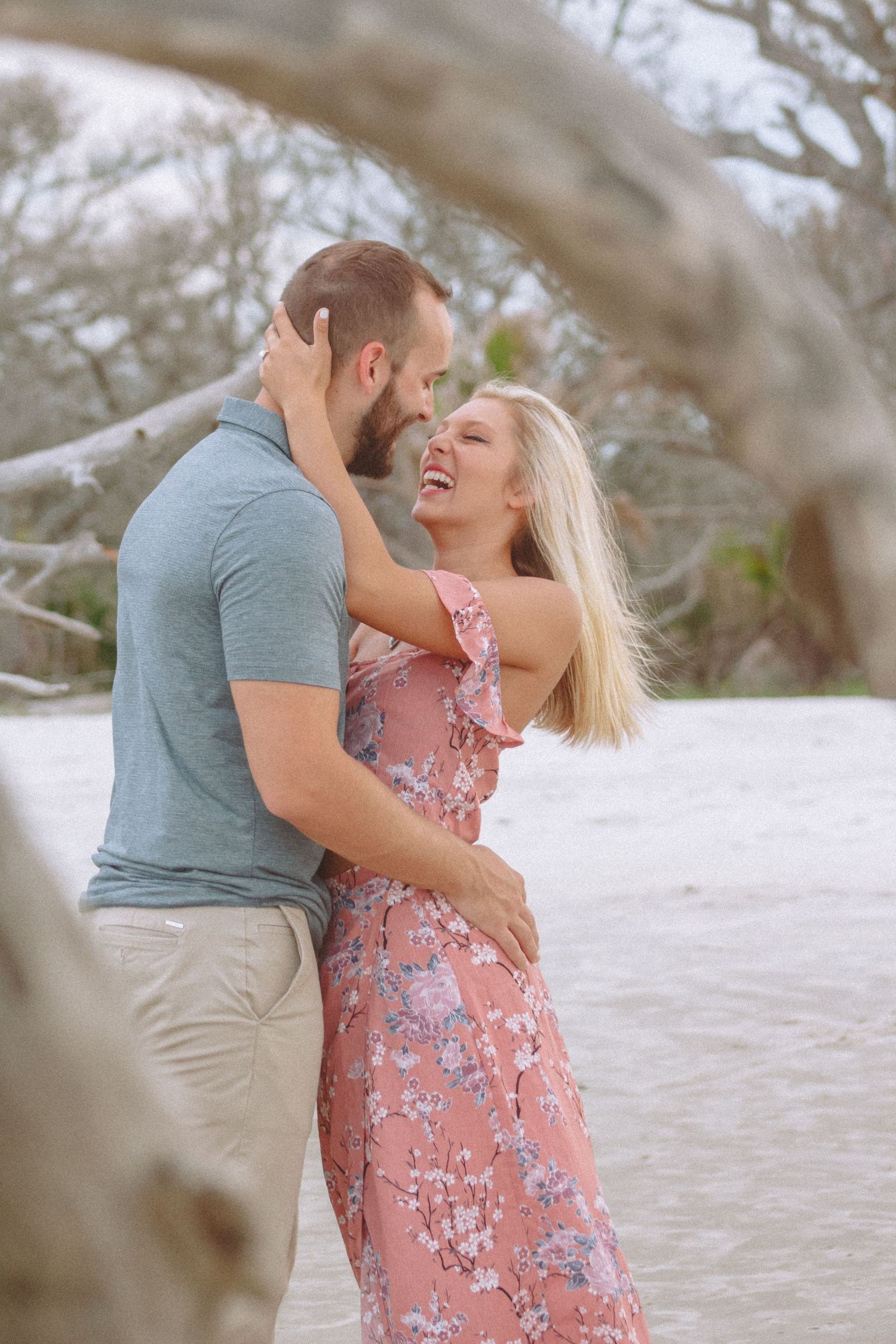 Laughing blonde woman in pink floral dress embracing bearded man on a white sand beach.