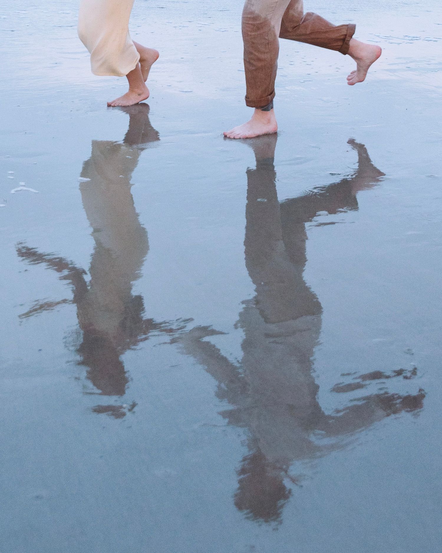 Barefoot couple walking on wet reflective sand at the beach, reflection visible below.