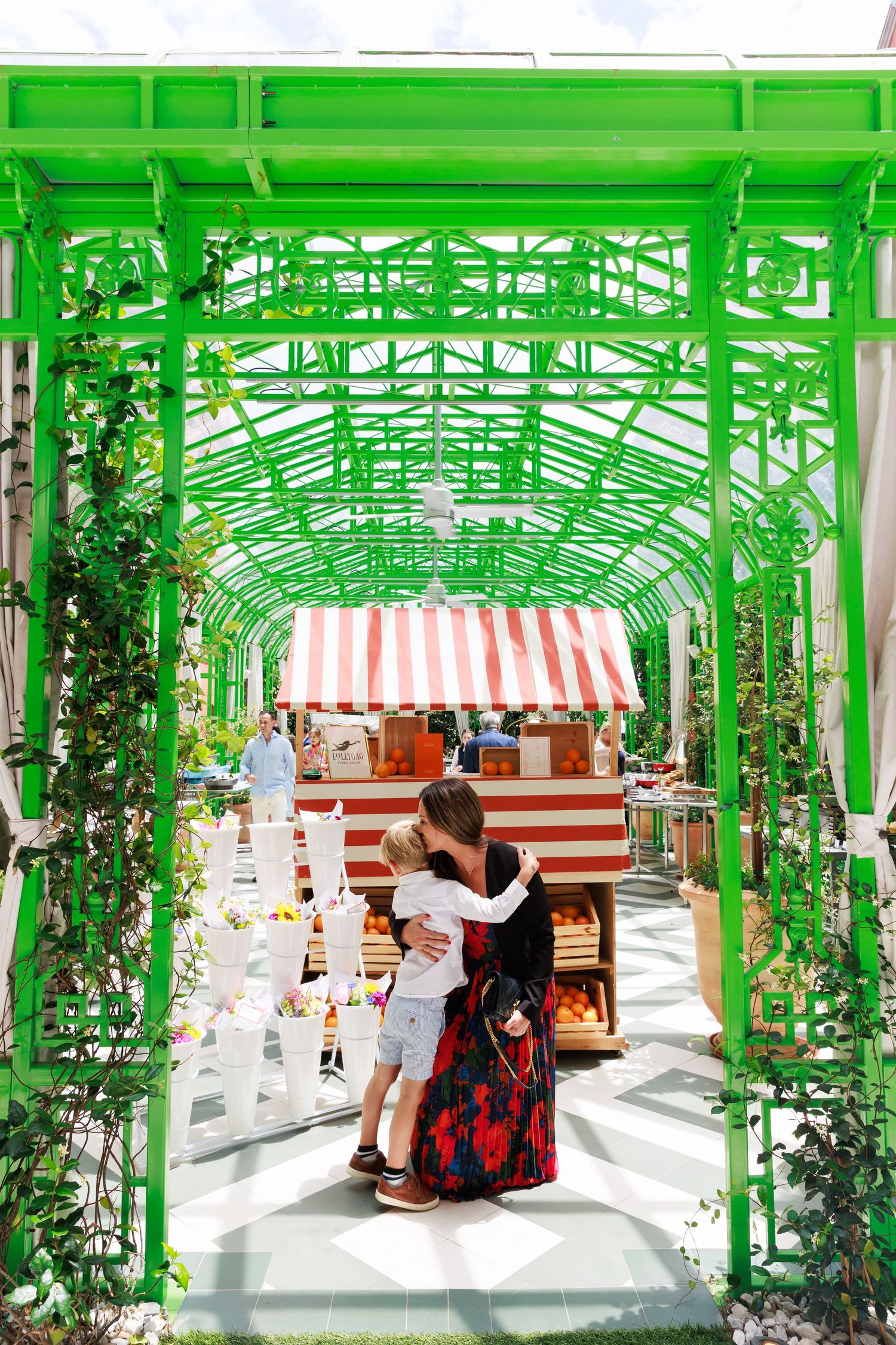 Woman in red dress hugging child under green metal pergola at outdoor market with striped canopy stall.