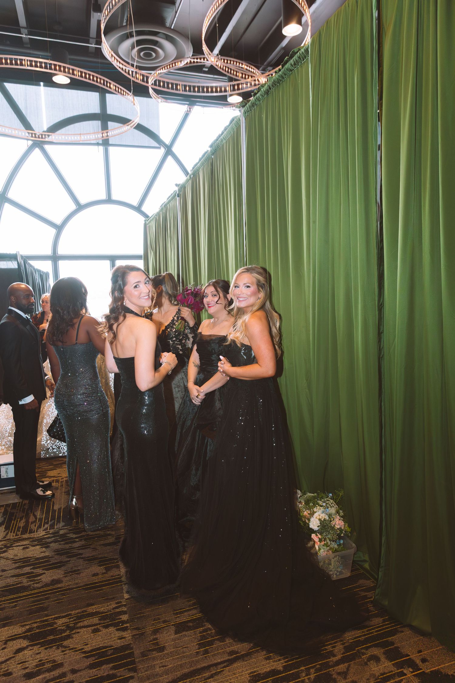 Bridesmaids in elegant black gowns gather near green curtains at a formal wedding reception venue.