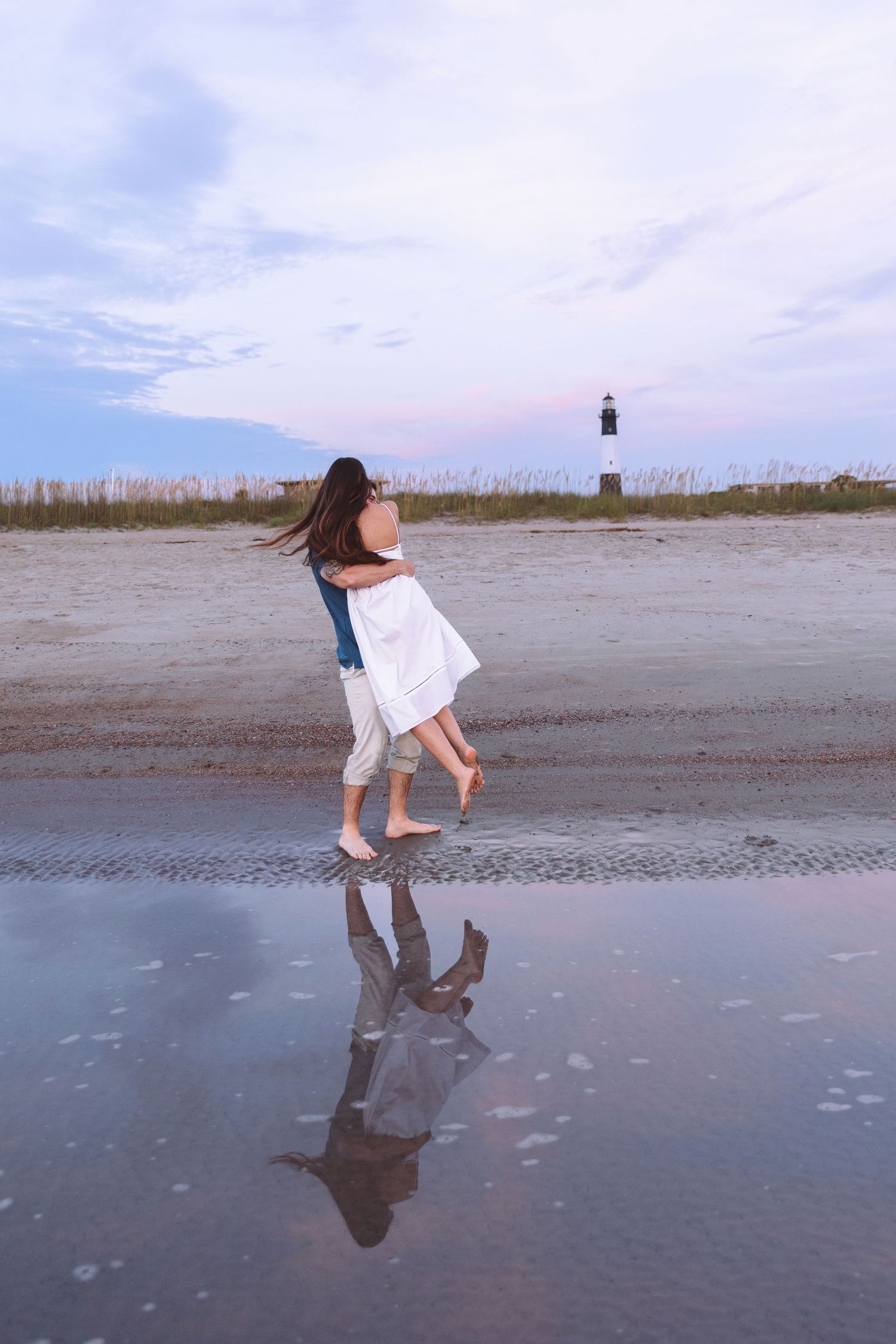 Couple dancing on a reflective wet beach at sunset with a lighthouse visible in the background.