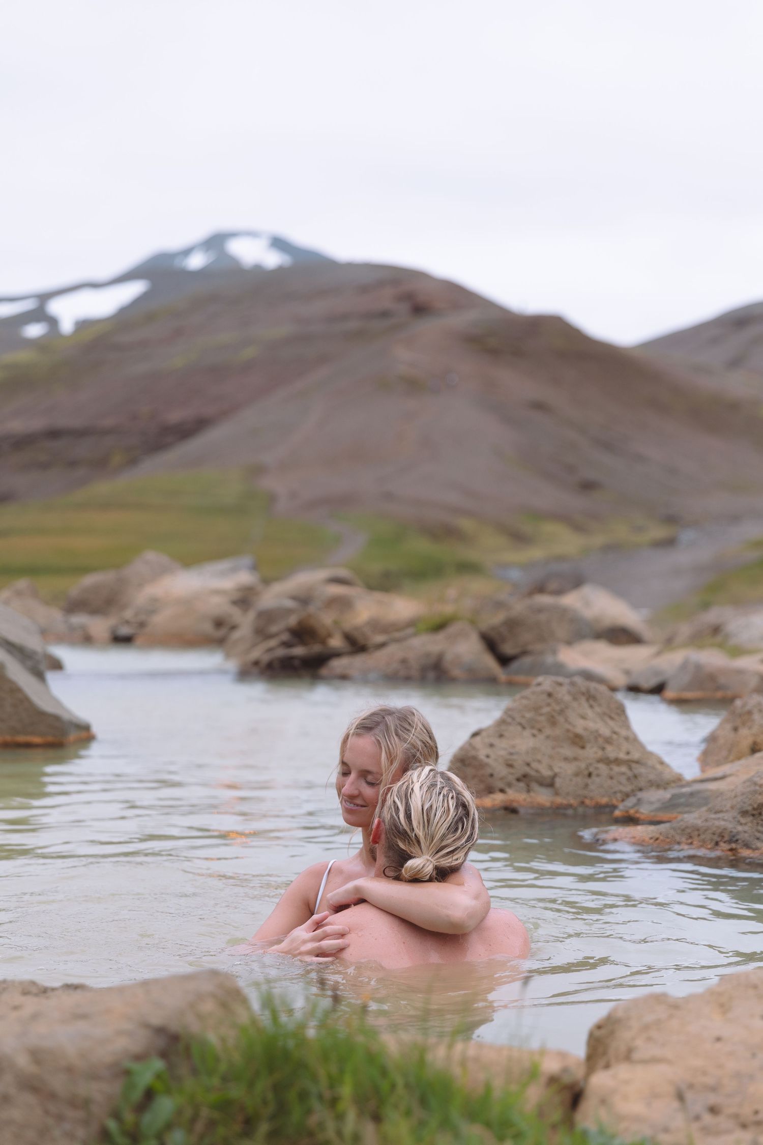 Couple embracing in a natural hot spring pool surrounded by rocks and mountains in Iceland.
