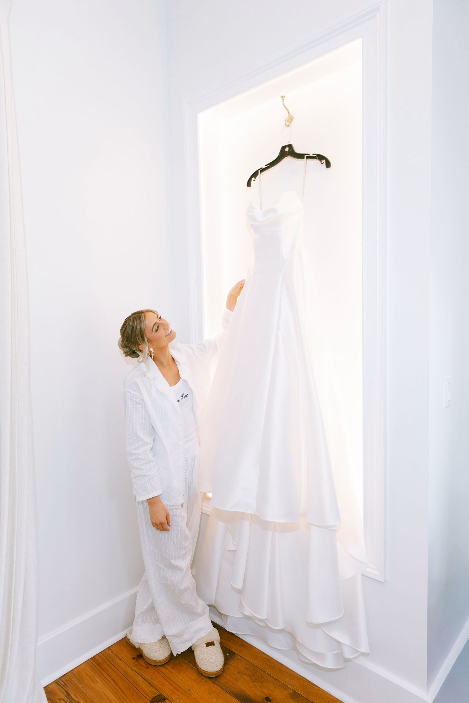 Smiling bride in white pajamas admiring her elegant strapless wedding gown hanging in a bright white room.