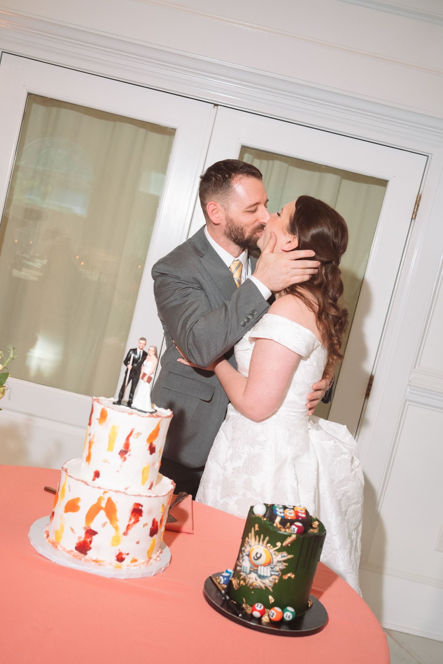Bride and groom kissing behind two-tier wedding cake and billiard groom cake on coral table.