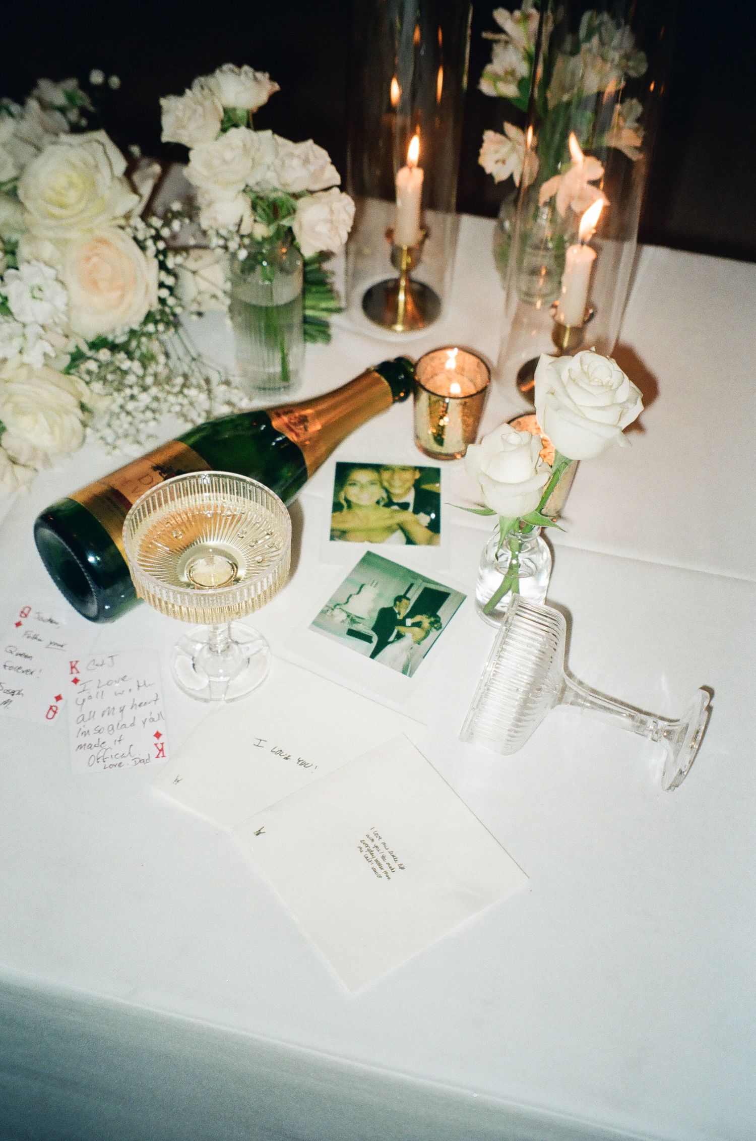Elegant wedding reception table with champagne bottle, Polaroid photos, white roses, candles, and handwritten notes on white linen.