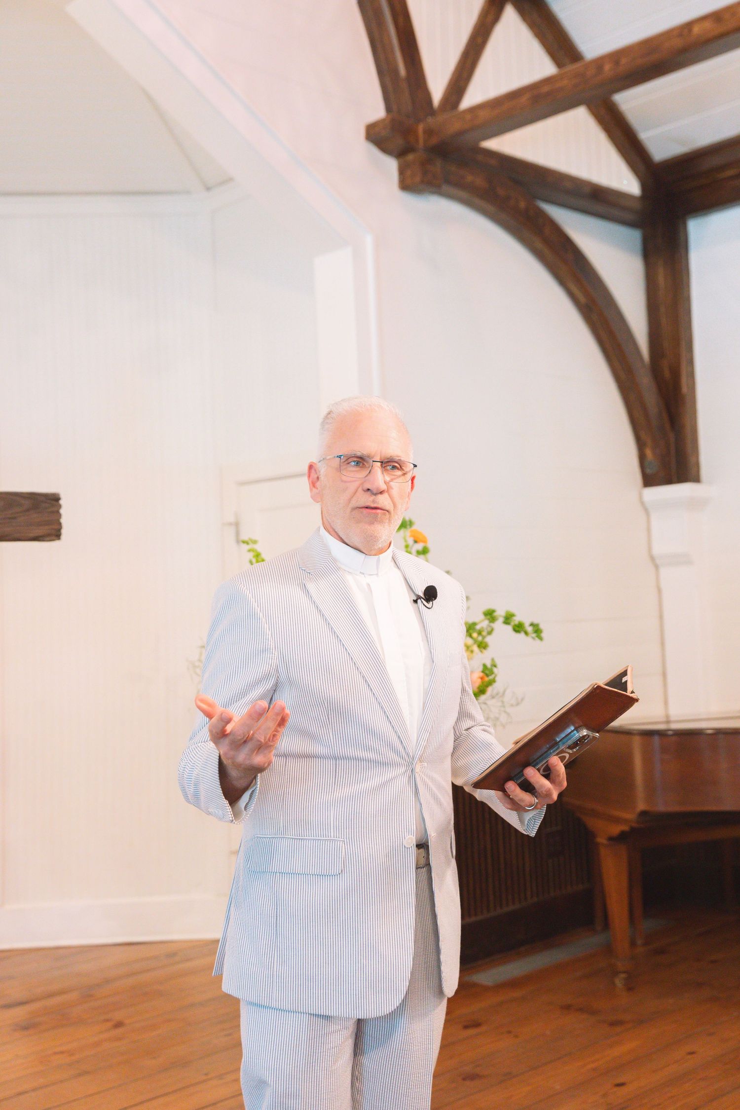 Elderly man in light gray suit holding book, speaking at indoor wedding ceremony with wooden beam ceiling.