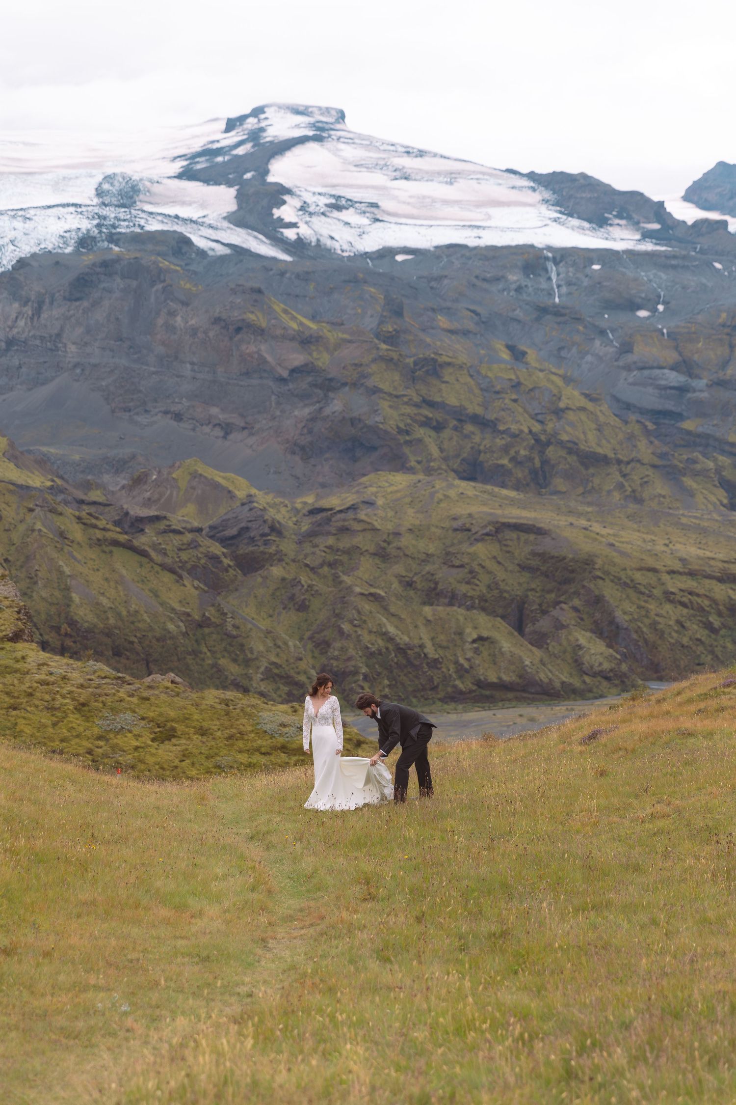 Wedding couple standing on green hillside with snow-capped Icelandic glacier and rugged mountains behind them.