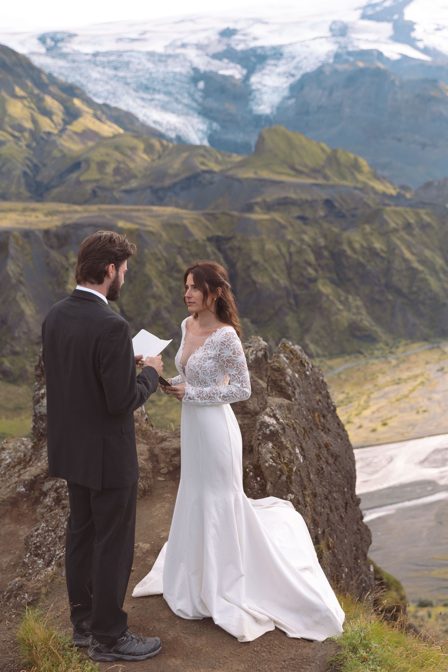 Couple reads wedding vows on dramatic Iceland highland cliff with glacier and river valley below.