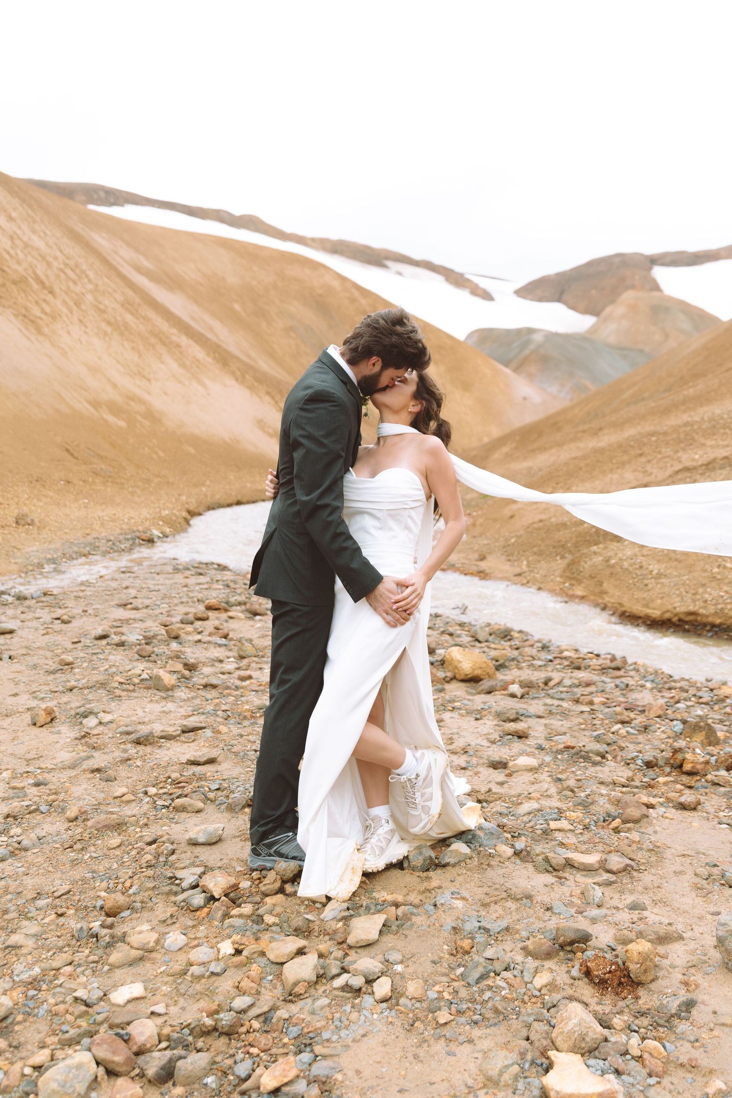 Groom kisses bride on cheek as she leans back in his arms on a rocky mountain landscape.