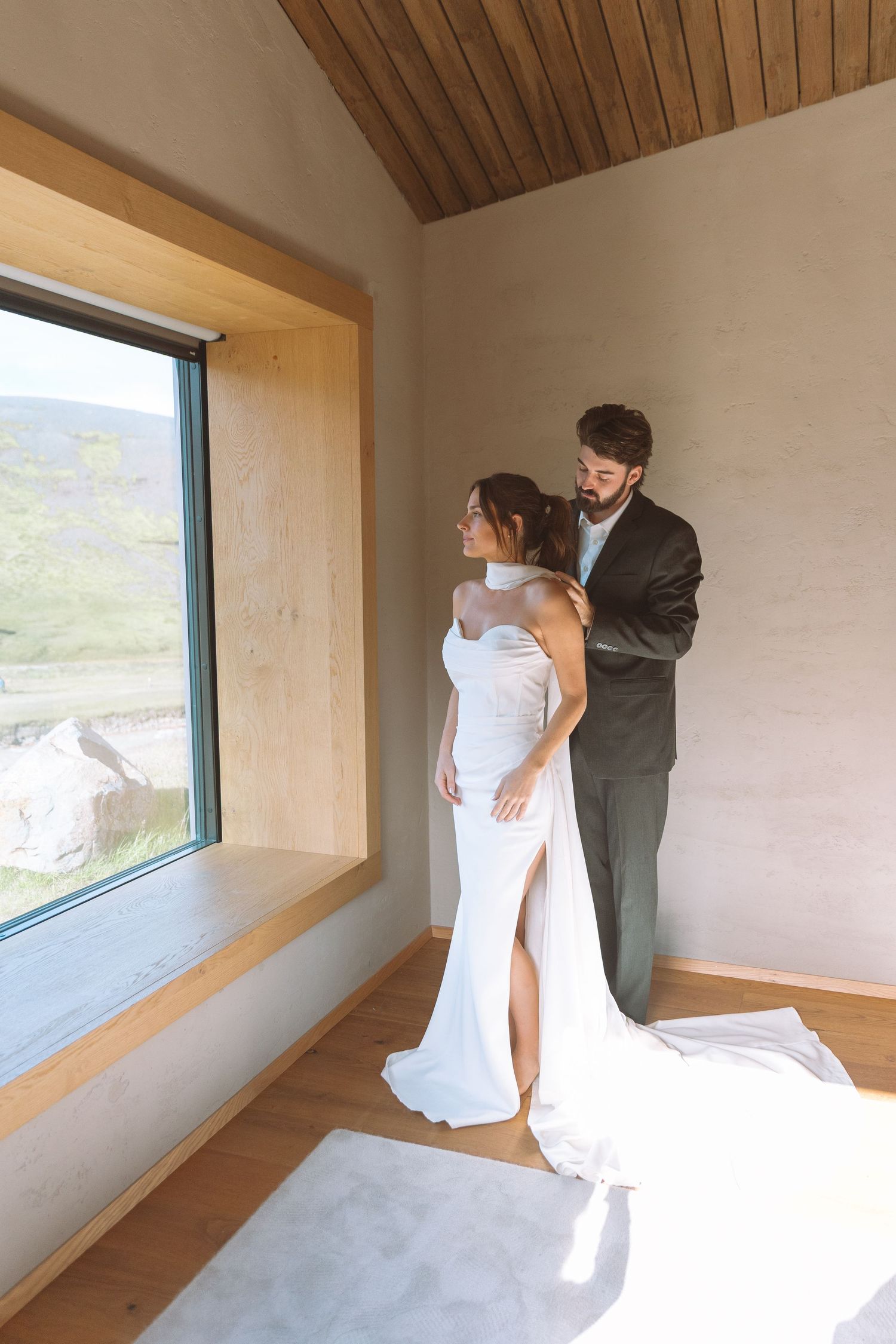 Groom lovingly adjusting brides white halter gown in a sunlit modern rustic room.