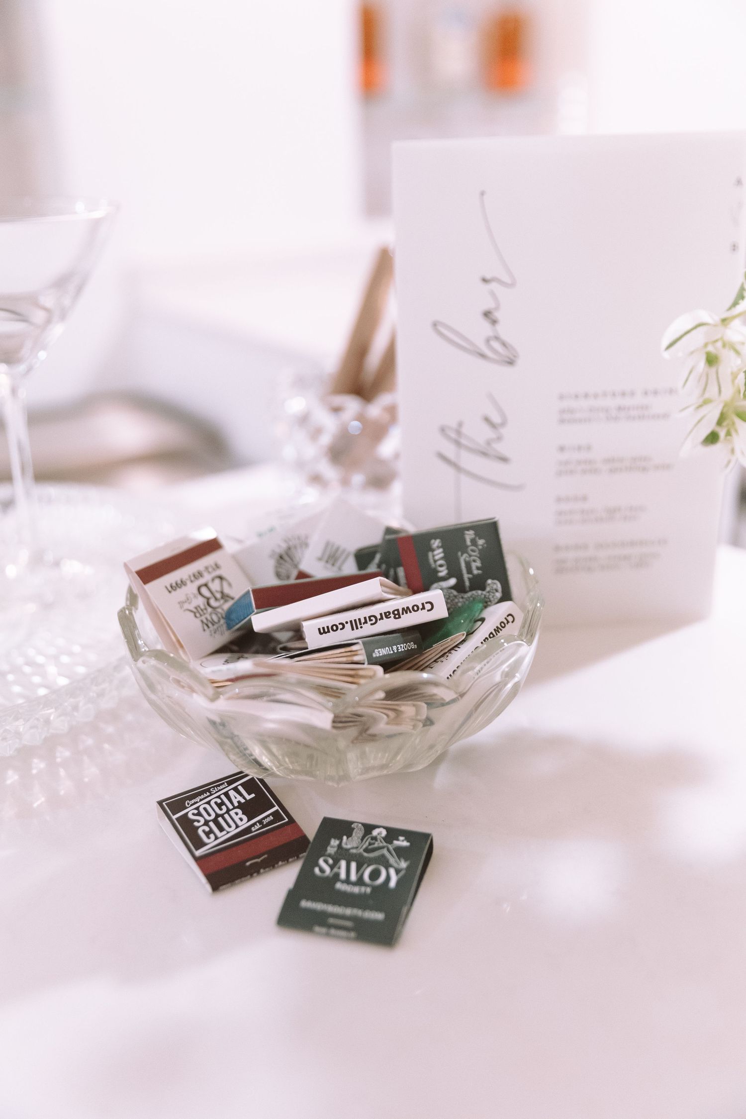 Glass bowl filled with matchbooks on a wedding bar table with elegant signage and crystal glassware.