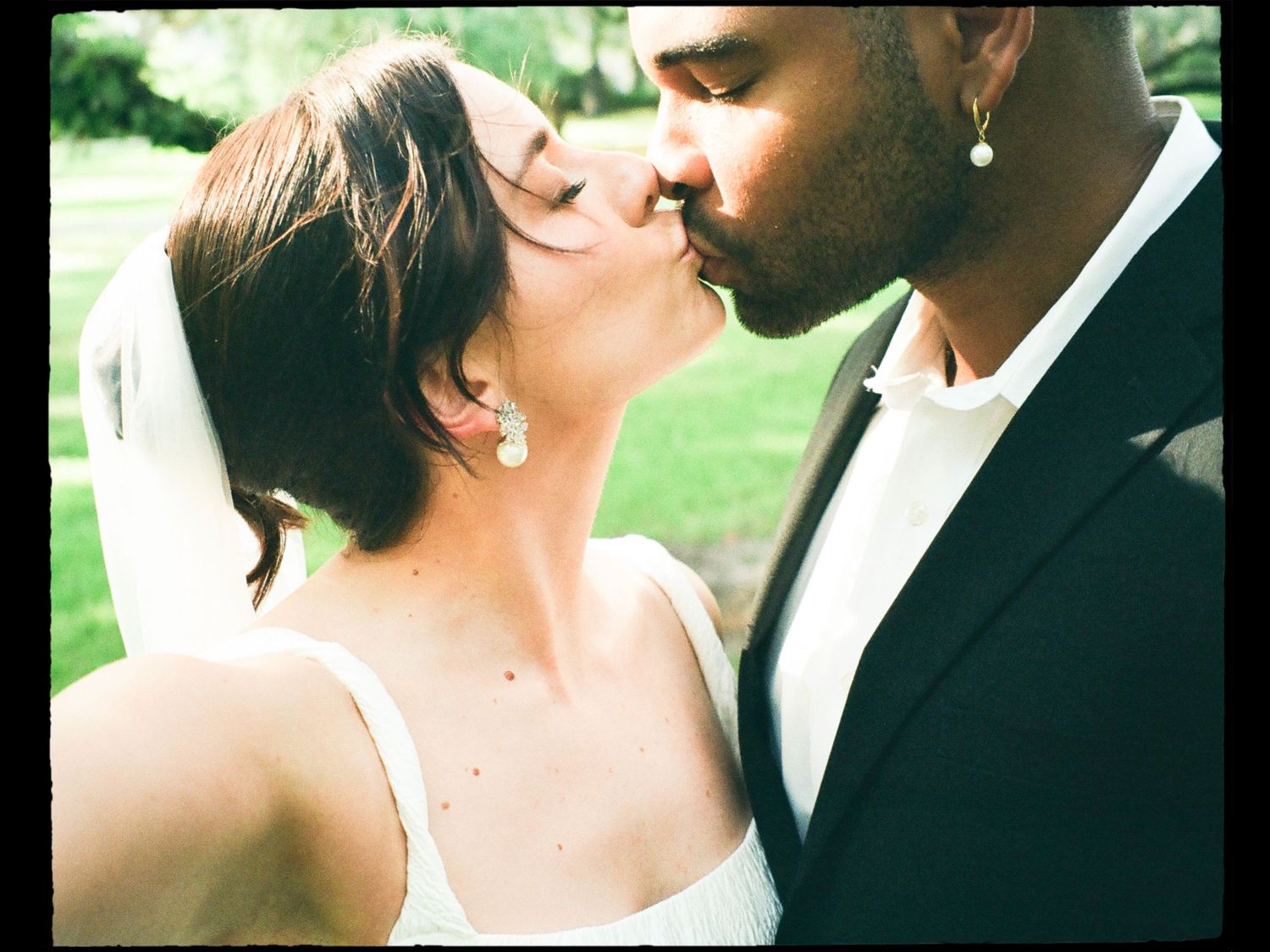 Bride and groom sharing a romantic kiss outdoors, with the bride wearing a veil and the groom in a black suit.