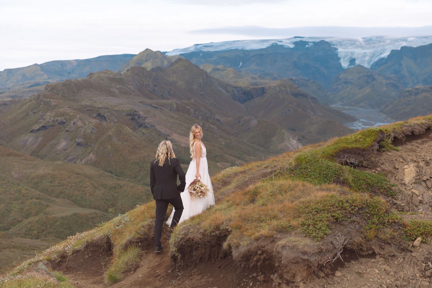 Bride and groom stand on a dramatic mountain ridge in Iceland, with sweeping valley and glacier views behind them.