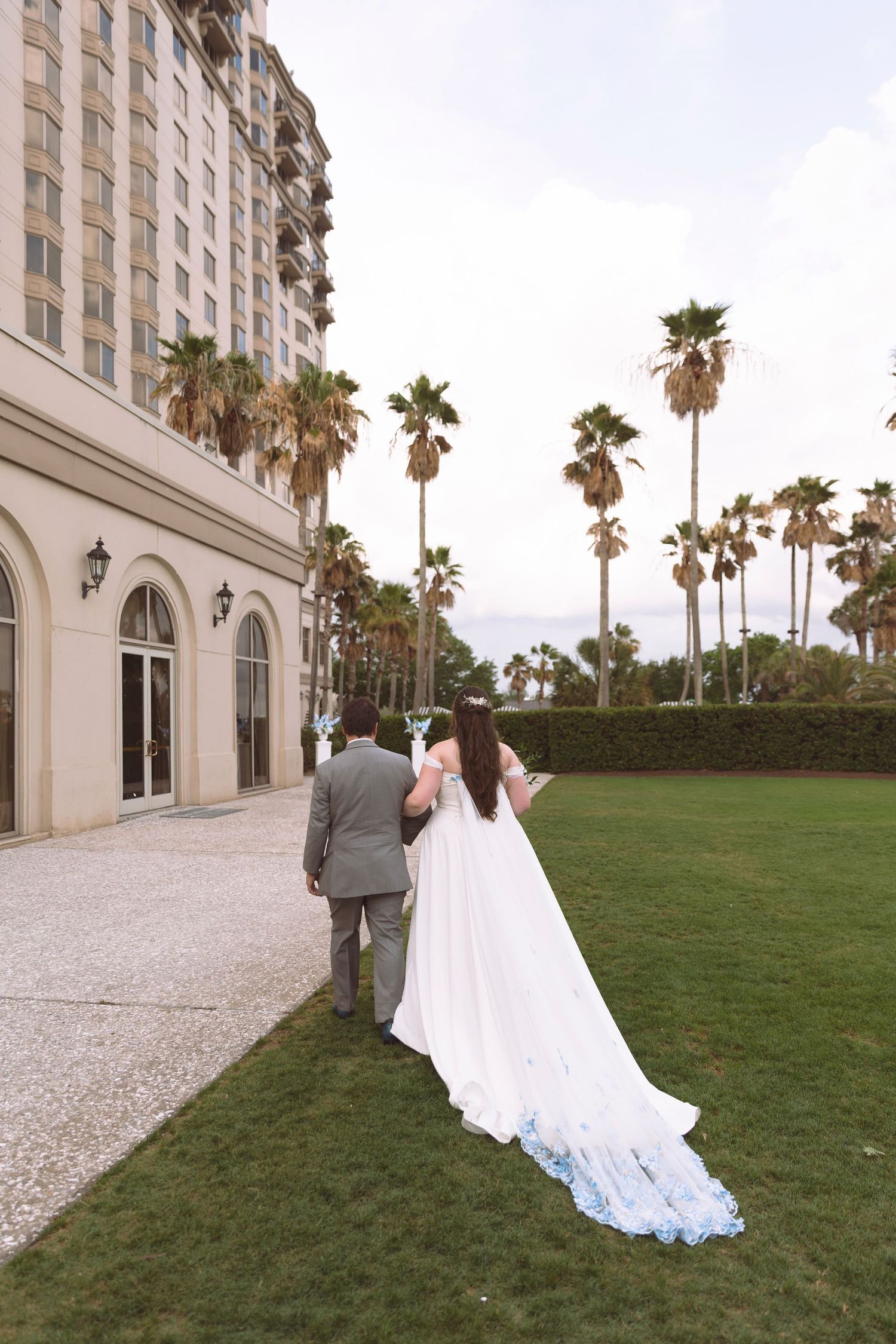 Bride in a white gown with blue-tipped train walks with groom across green lawn beside a luxury hotel with palm trees.