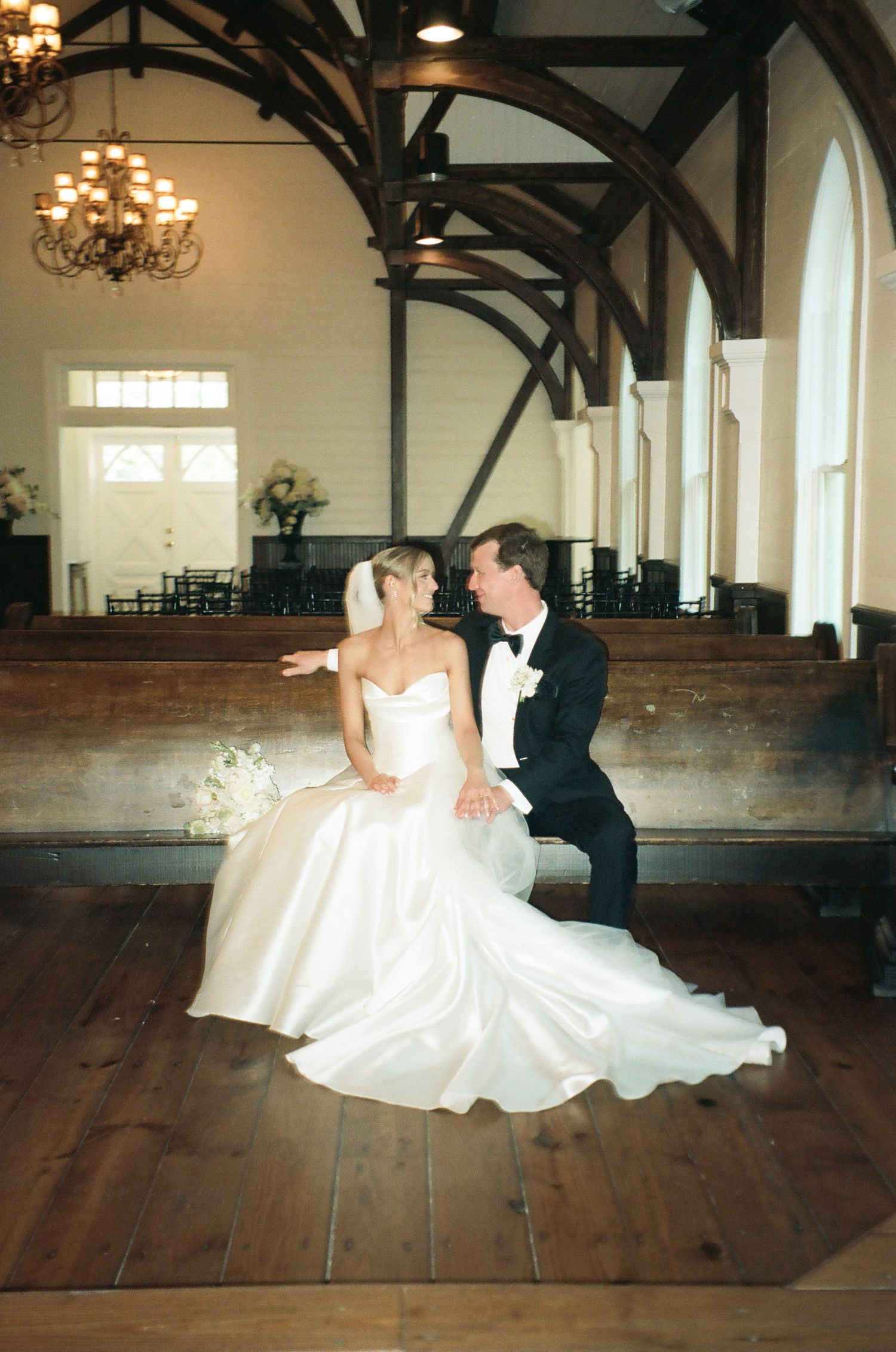 Bride and groom sit together on a wooden pew in a white chapel, sharing a romantic moment after their wedding.