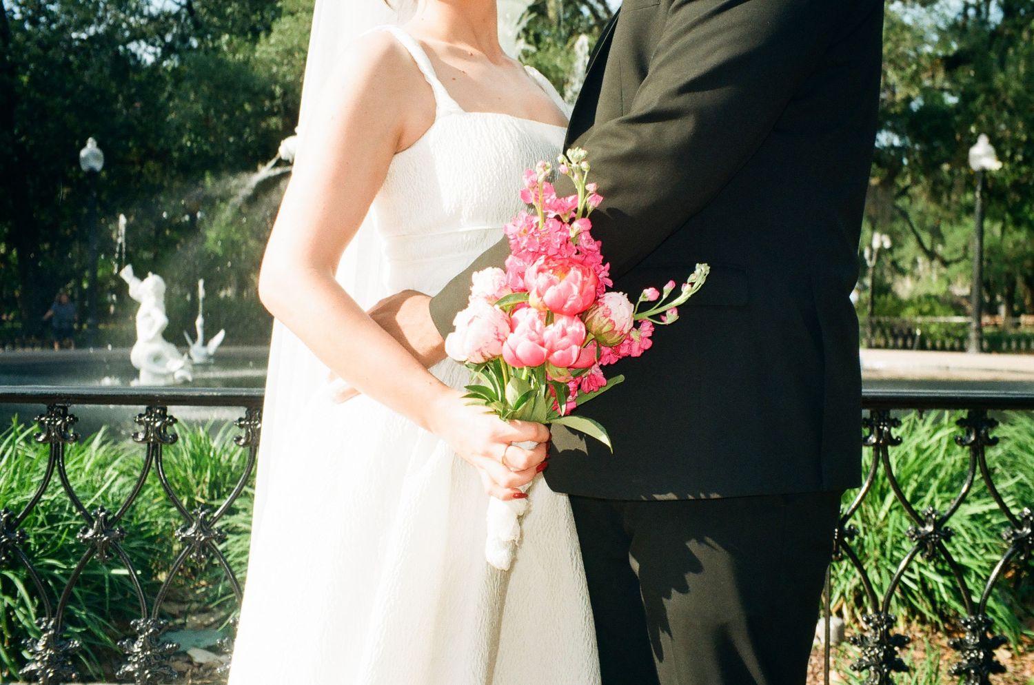 Bride in white gown holding pink floral bouquet stands beside groom in black suit near ornate iron fence and fountain.
