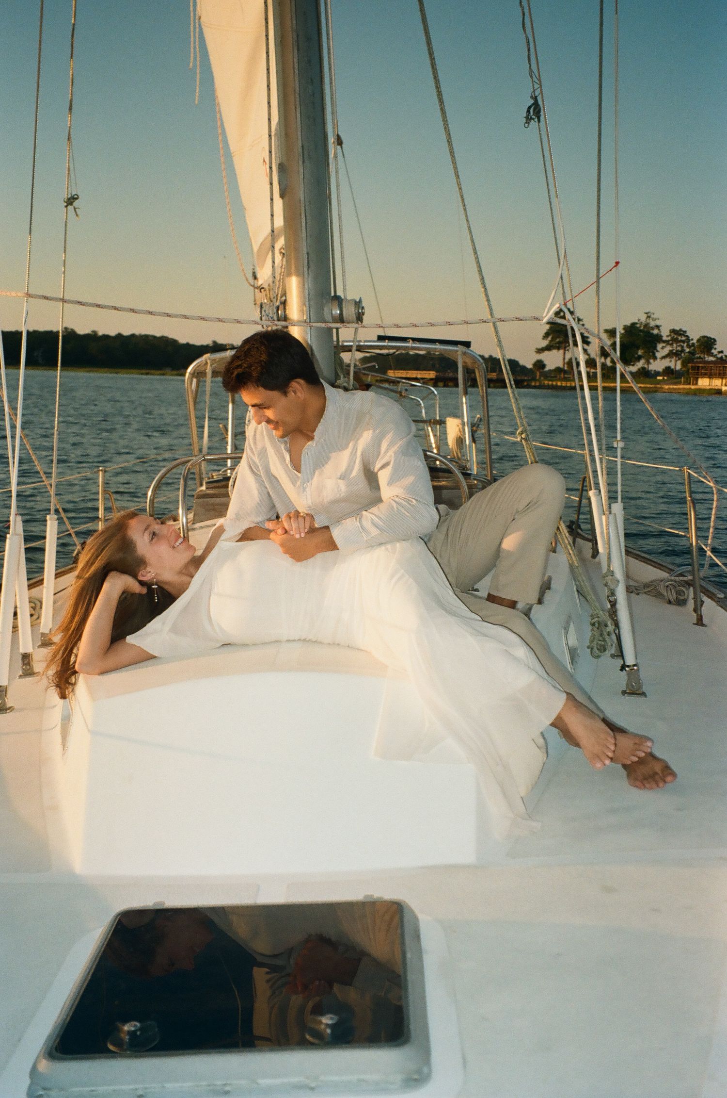 Couple in white outfits sharing a romantic moment on a sailboat at golden hour sunset.