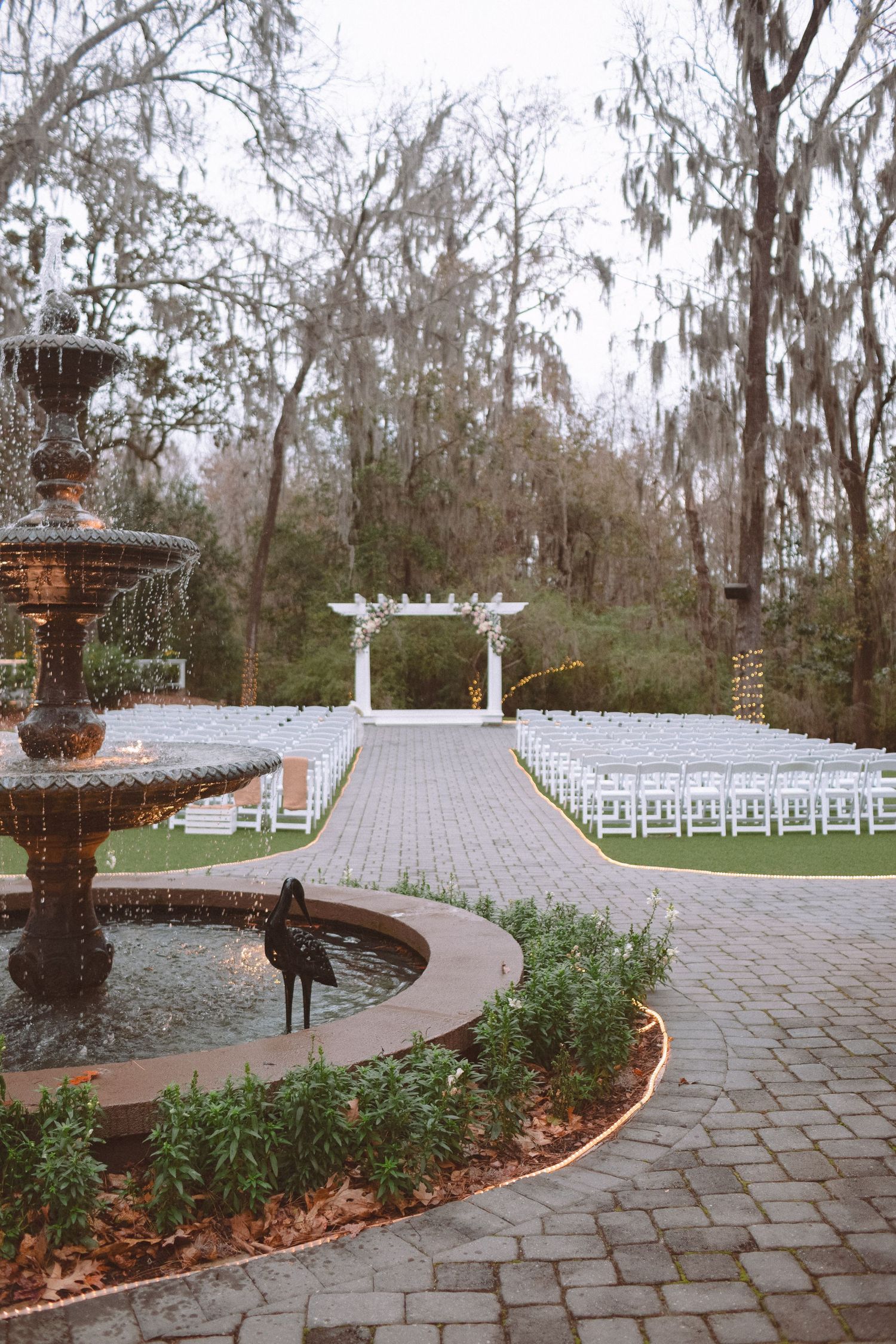 Outdoor wedding ceremony setup with white chairs, pergola altar, fountain, and Spanish moss-draped trees.