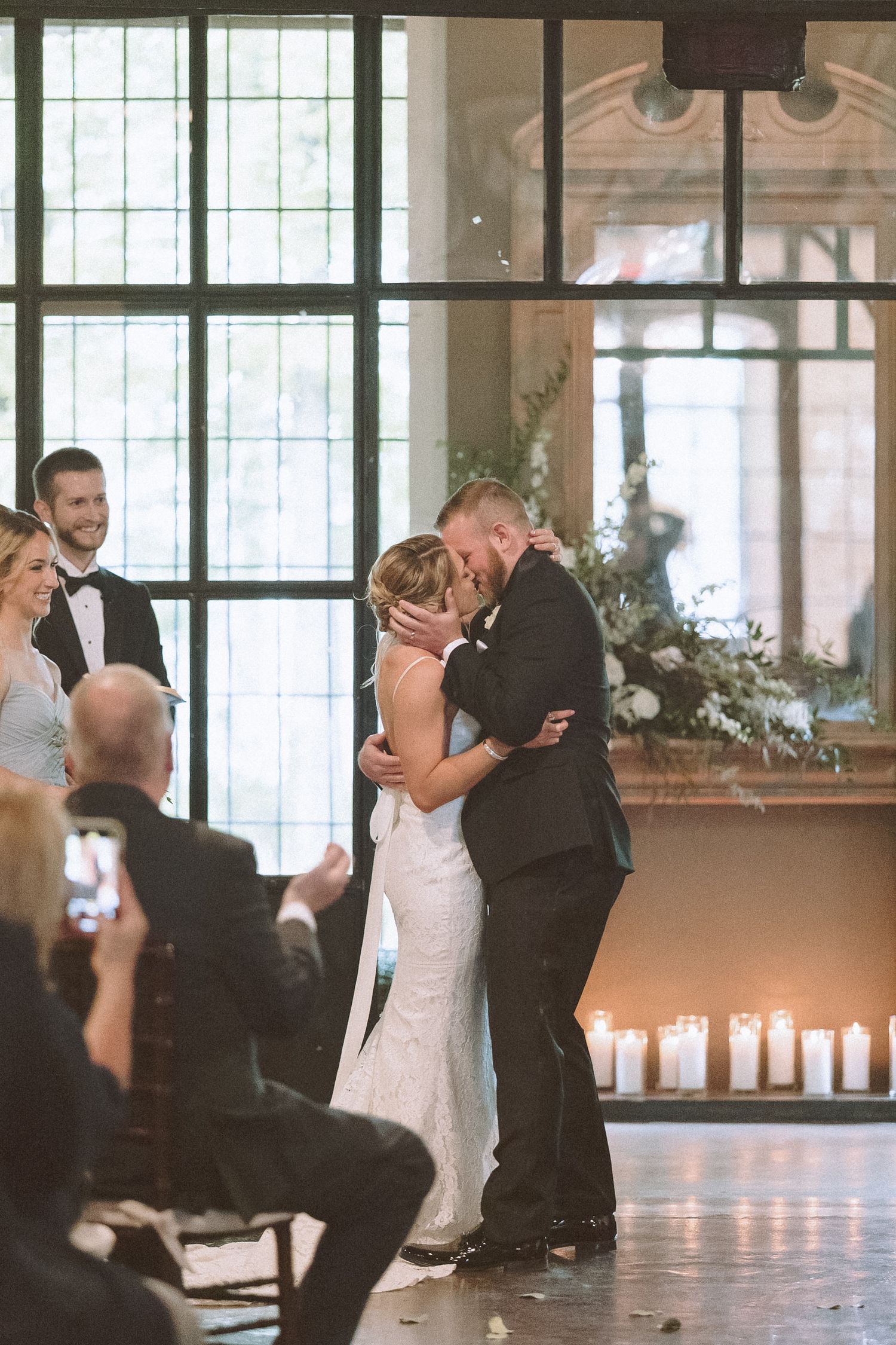 Bride and groom share first kiss at altar surrounded by candles and floral arrangements.