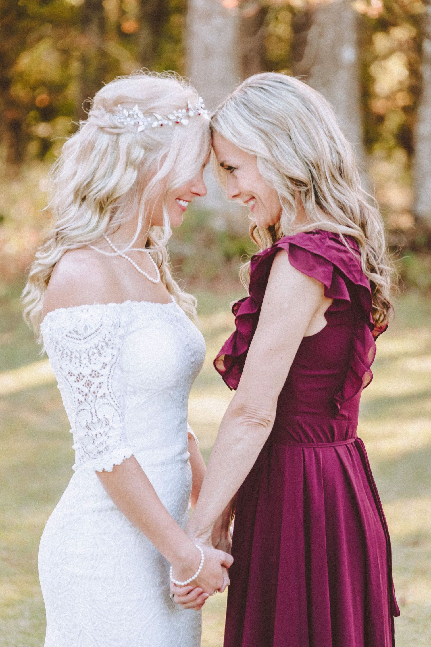 Two blonde women in wedding attire holding hands and smiling at each other outdoors.