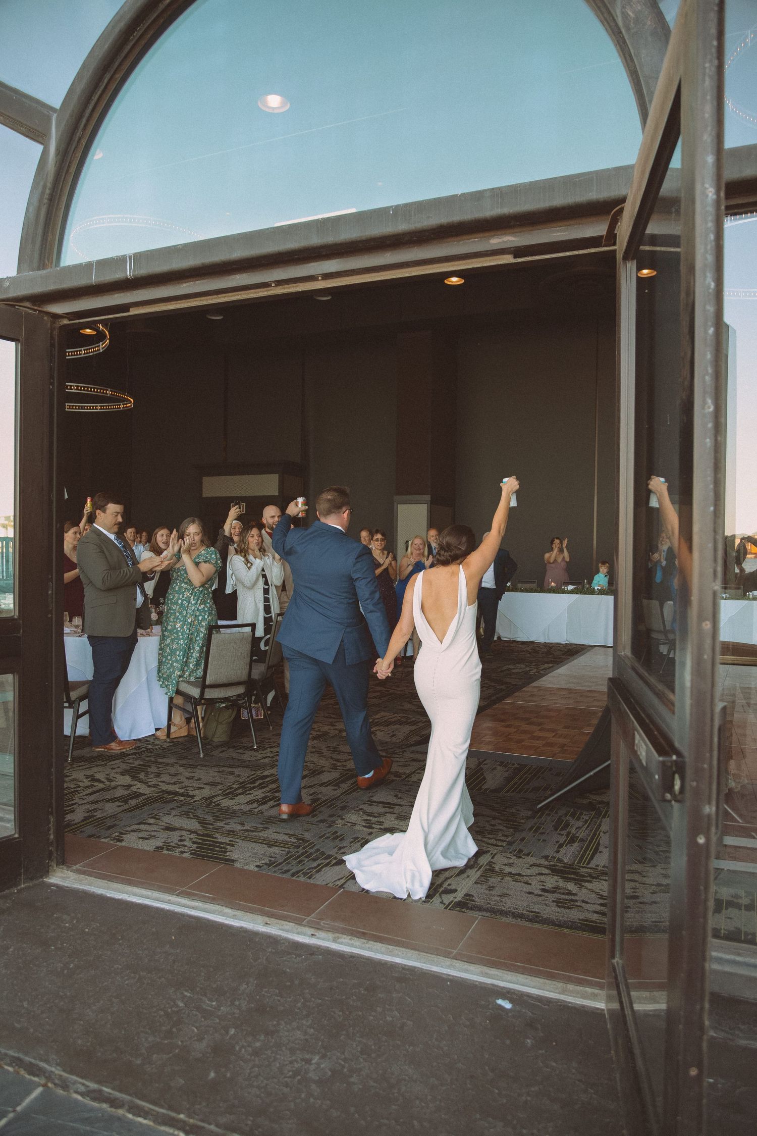 Bride and groom make grand entrance into wedding reception hall as guests cheer and applaud.