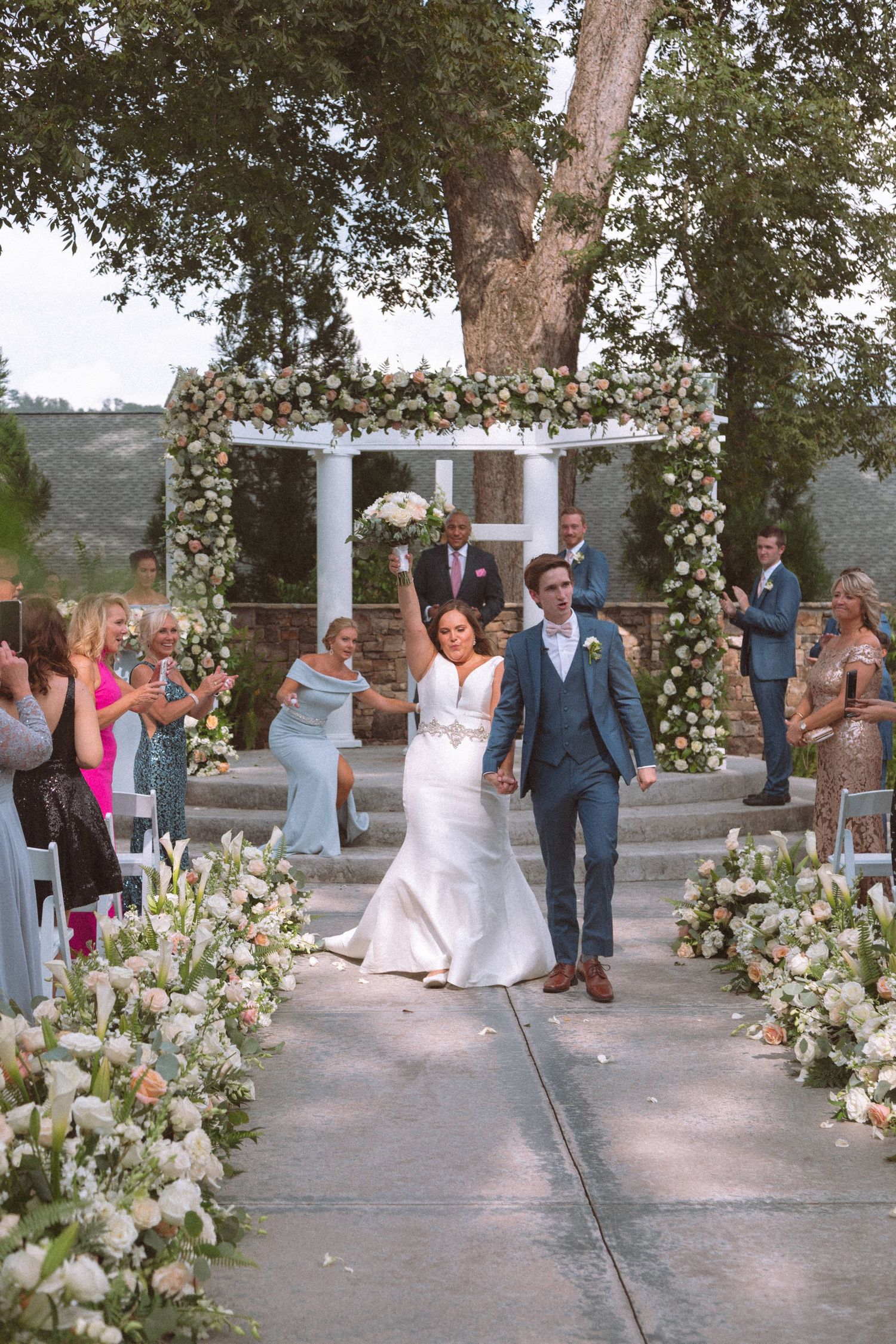 Joyful bride raises bouquet while walking down aisle with groom under floral arch after ceremony.
