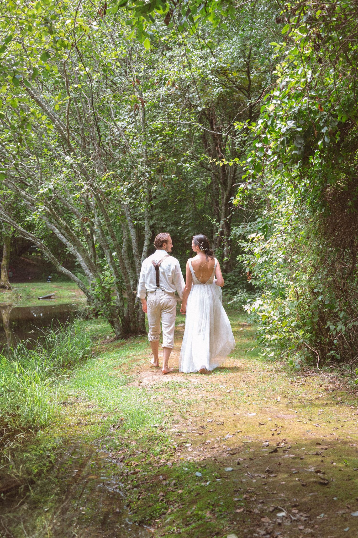 Bride and groom walking hand in hand barefoot down a lush green forest path.