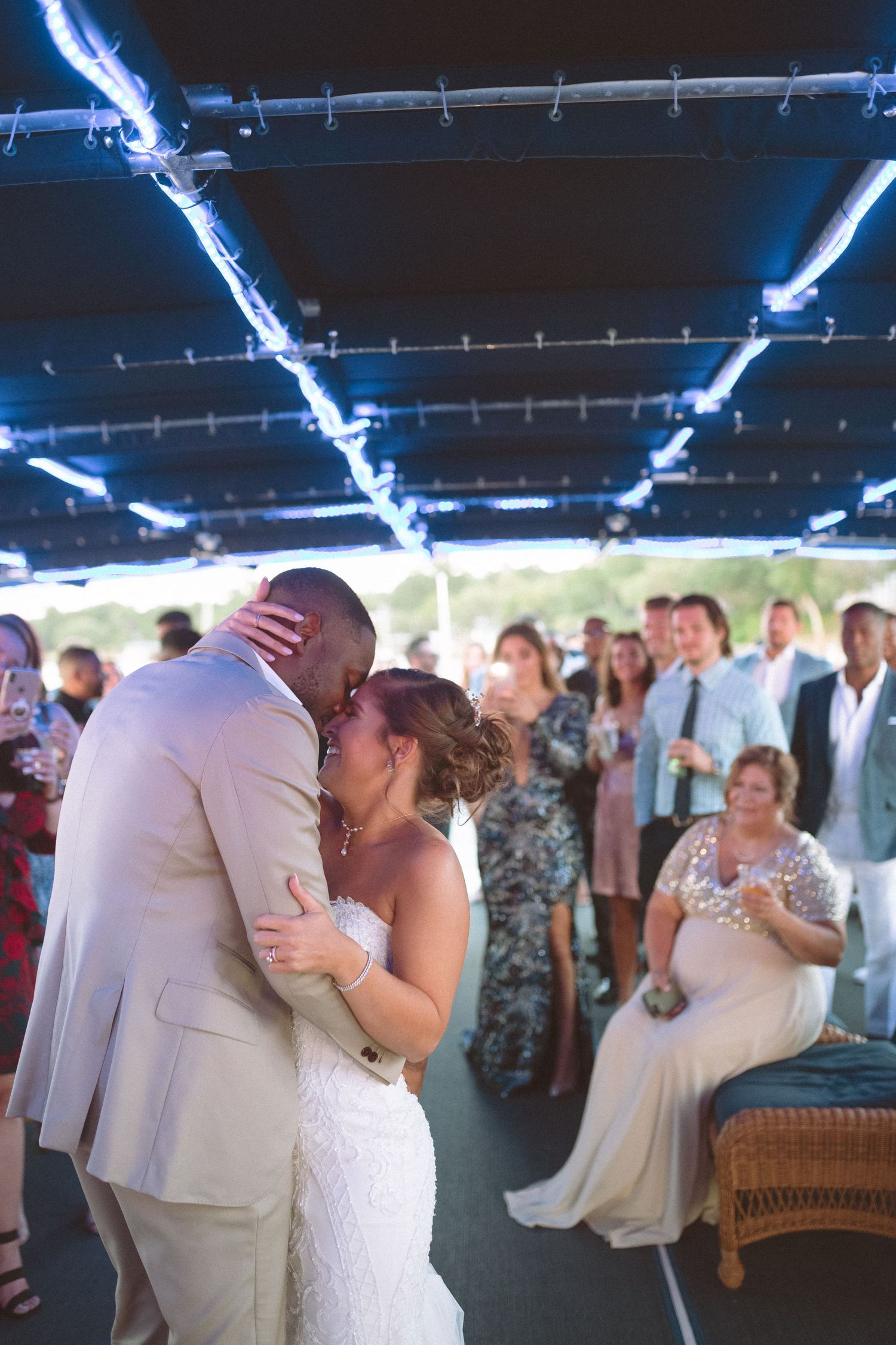 Newlyweds sharing their first dance kiss surrounded by guests on a boat wedding reception deck.