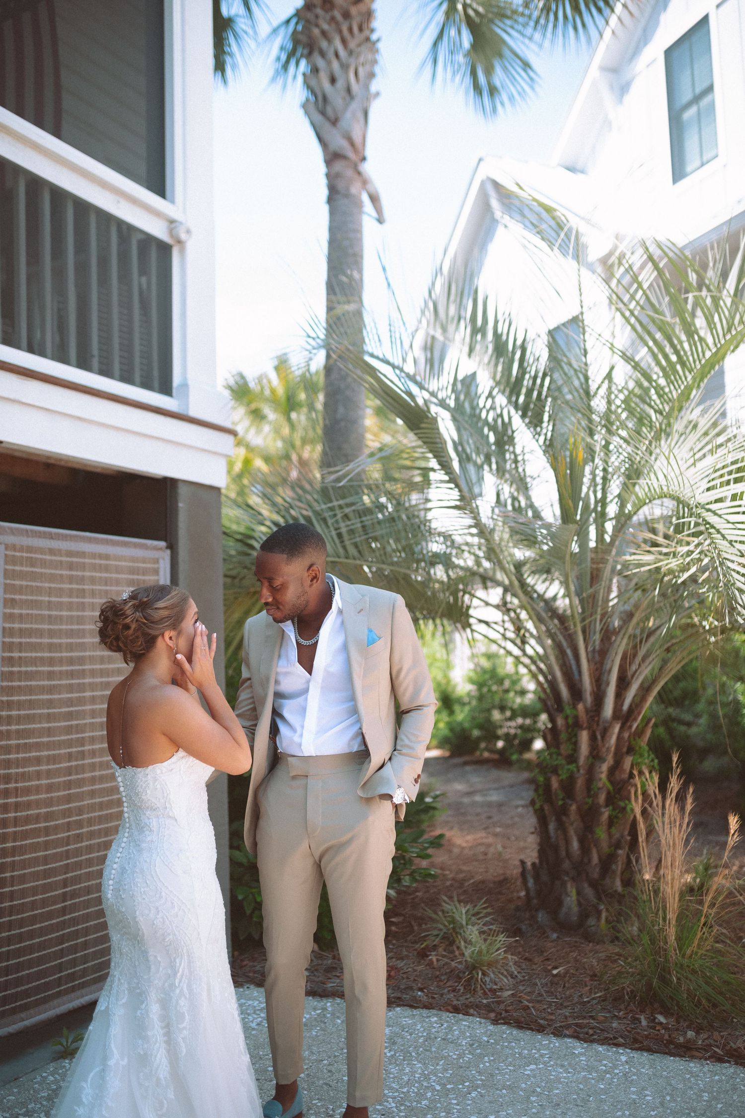 Bride wipes tears during intimate first look moment with groom in tan suit beside tropical palm trees.