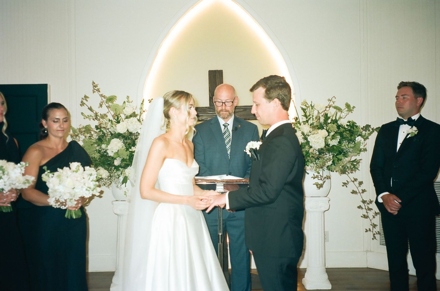 Smiling bride and groom exchange vows at elegant indoor ceremony with white floral arrangements and officiant.