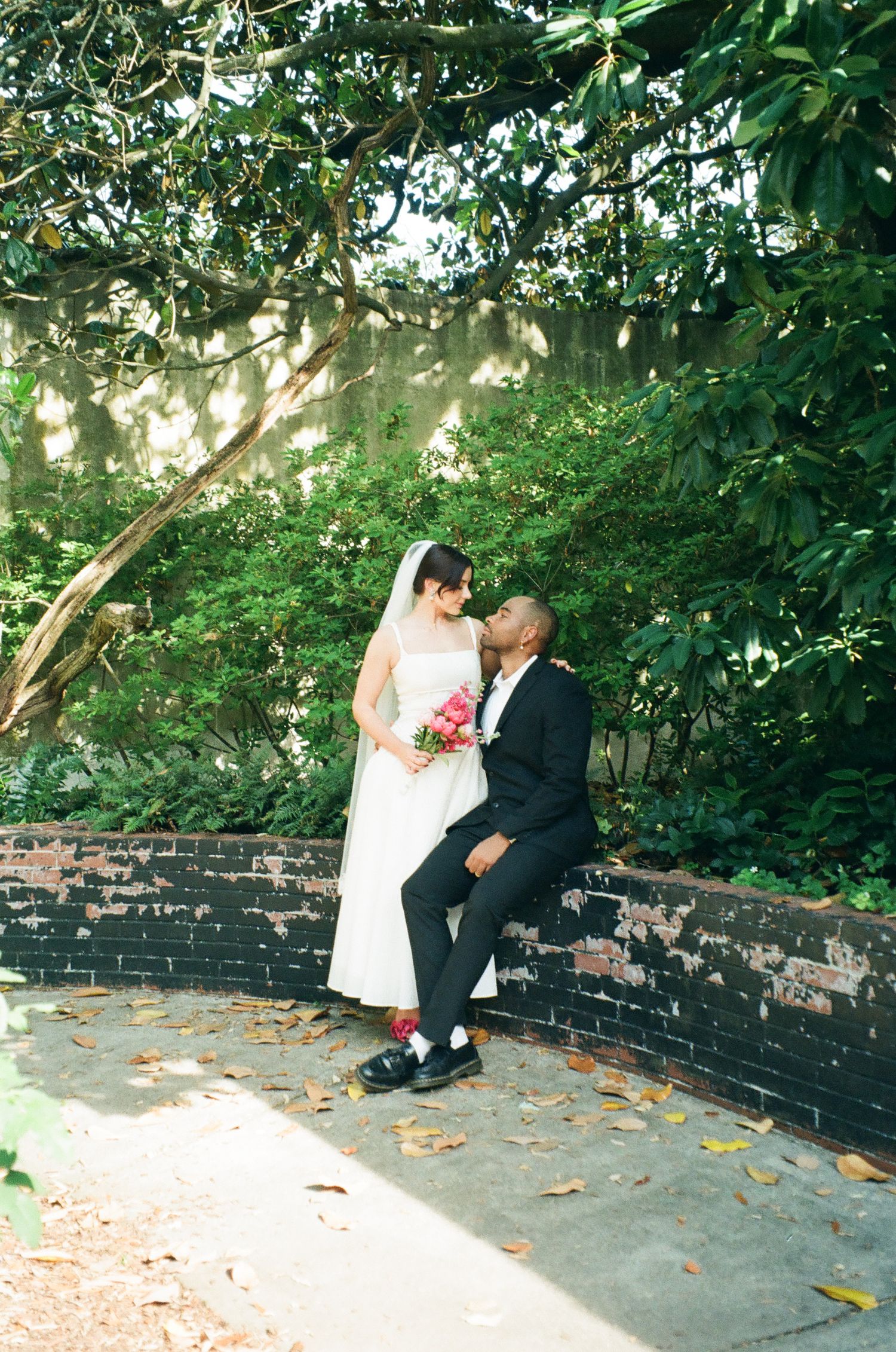 Bride and groom share a romantic moment in a lush garden, bride holding a pink bouquet in a white gown and veil.