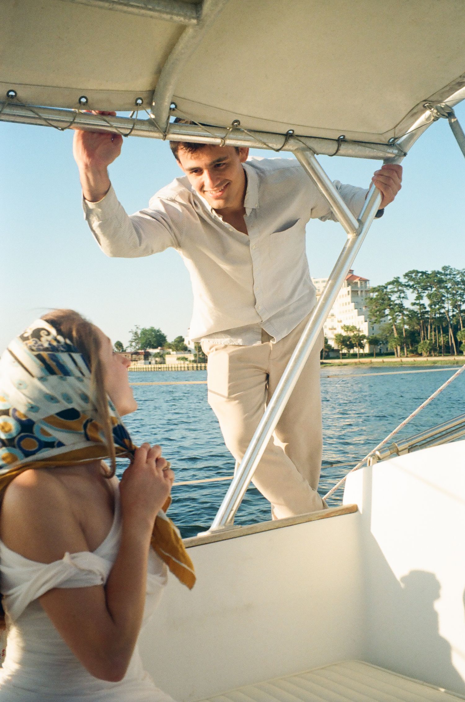 A smiling man in white linen holds a boat canopy while a woman in a headscarf sits on a sunny waterway.