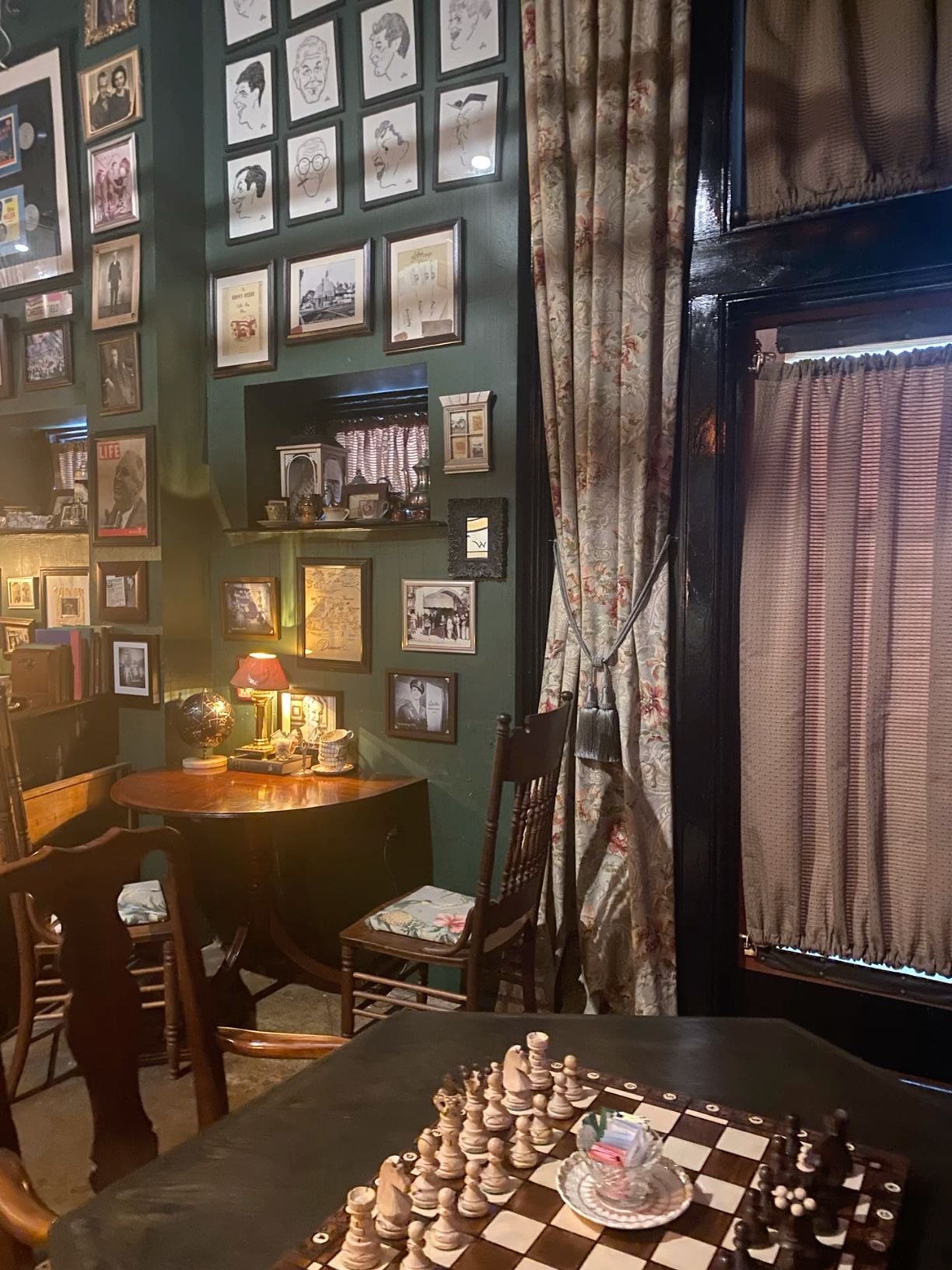 Cozy vintage bar corner with chess board, framed photos on dark green walls, and elegant curtains.