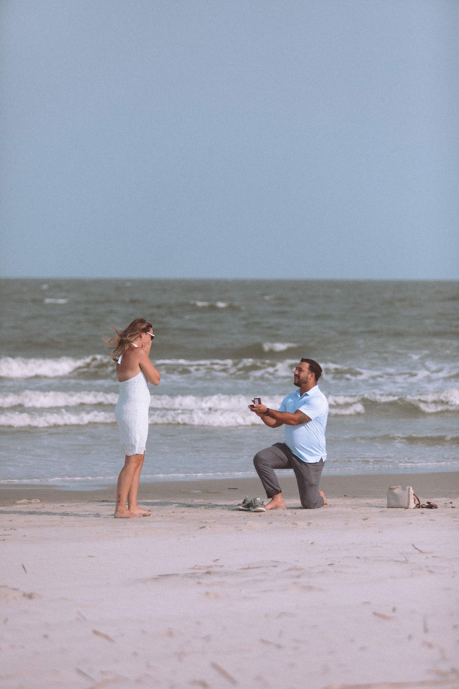 Man proposing on one knee to woman in white dress on a sunny ocean beach.