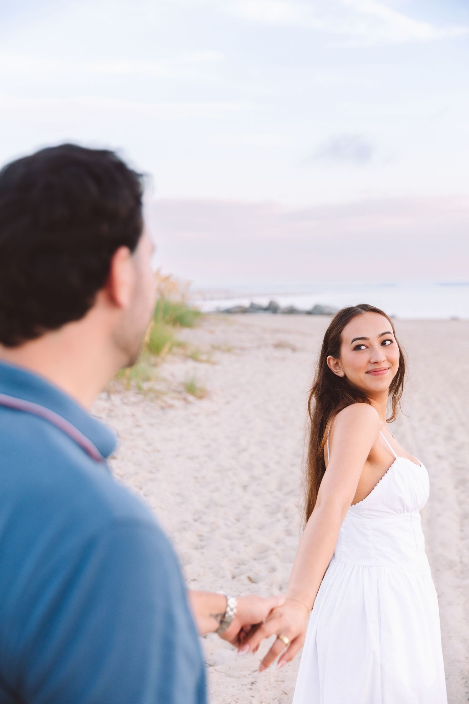 Couple holding hands on a sandy beach at sunset, woman in white dress smiling back.