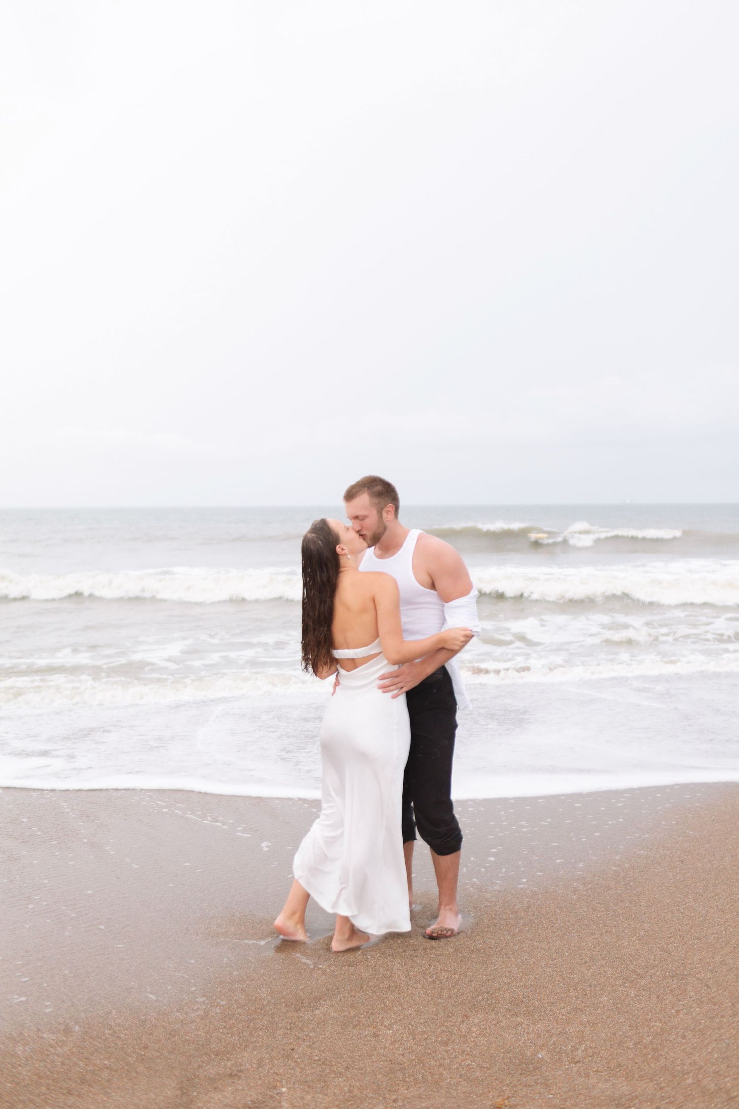 Couple sharing a tender kiss while standing barefoot on a beach with ocean waves in background.