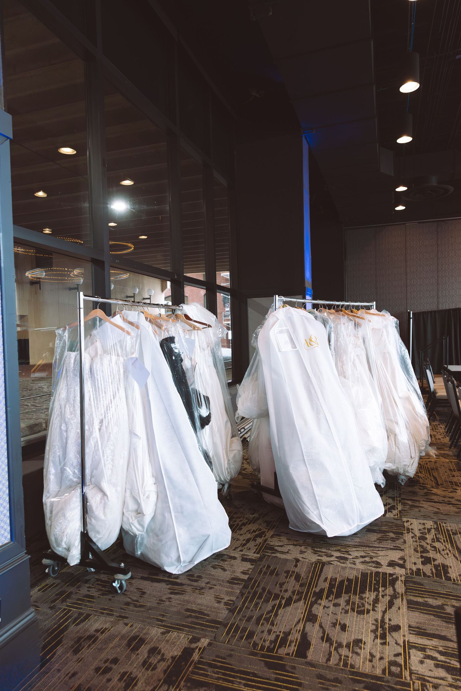 Wedding gowns in plastic covers displayed on rolling racks in a ballroom venue corridor.