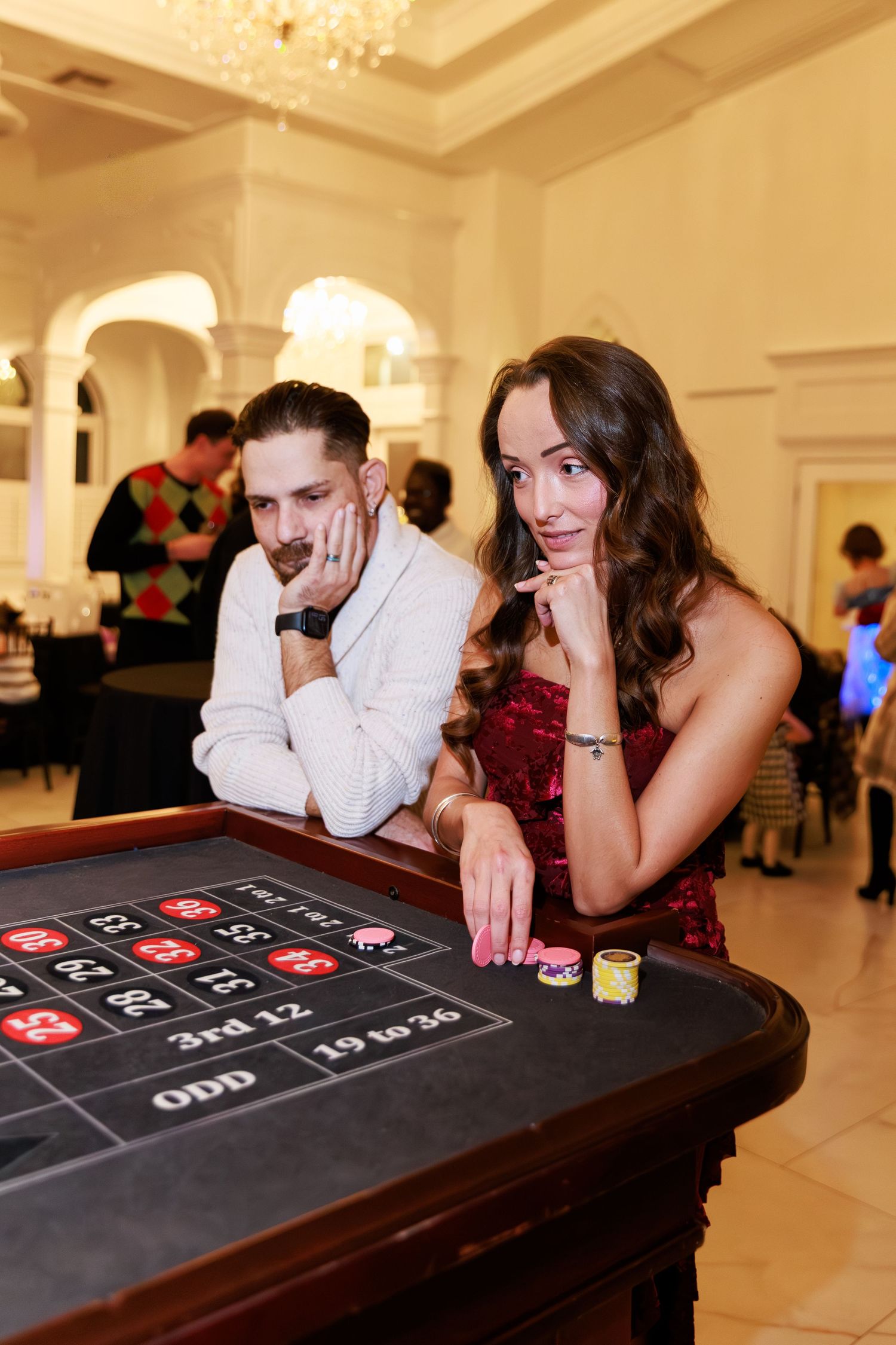 A couple poses at a roulette table with casino chips during an elegant event in a grand ballroom.